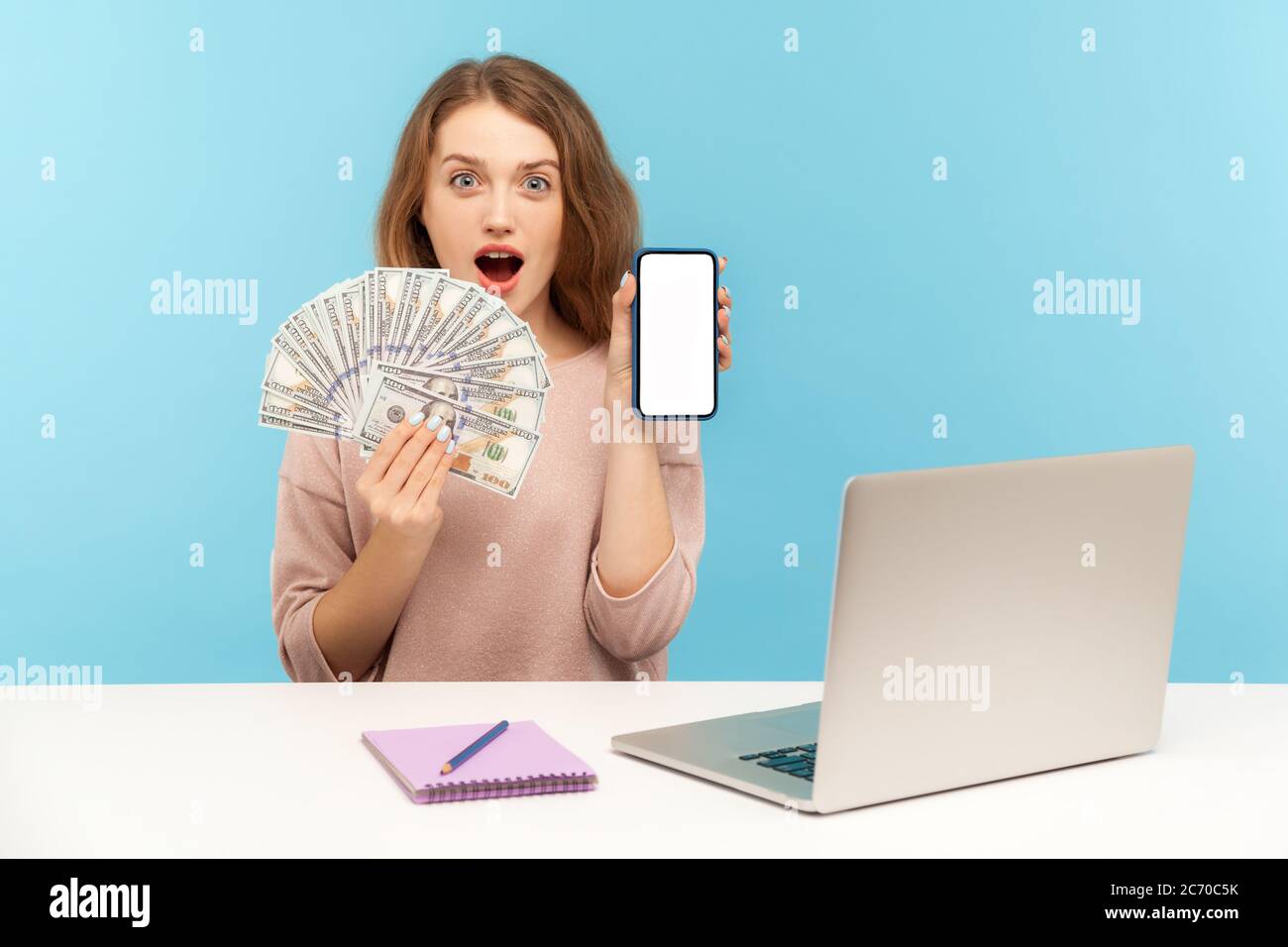 Internet earnings, online payment. Amazed woman freelancer sitting at workplace and holding dollar banknotes, showing smartphone with mockup display, Stock Photo