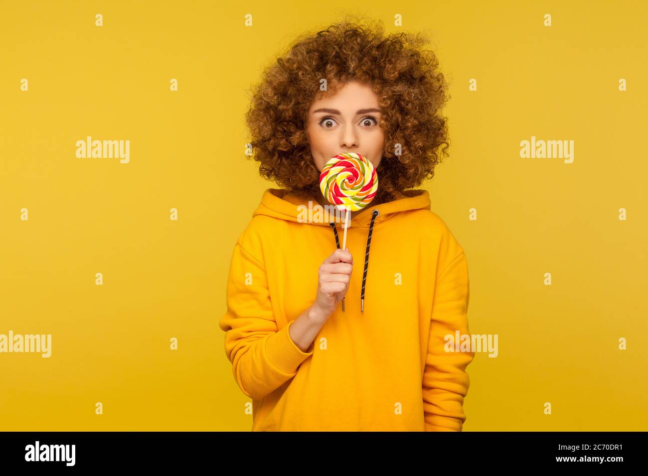 Sweet rainbow candy. Portrait of positive modern curly-haired woman in urban style hoodie licking lollipop and looking at camera with amazed expressio Stock Photo