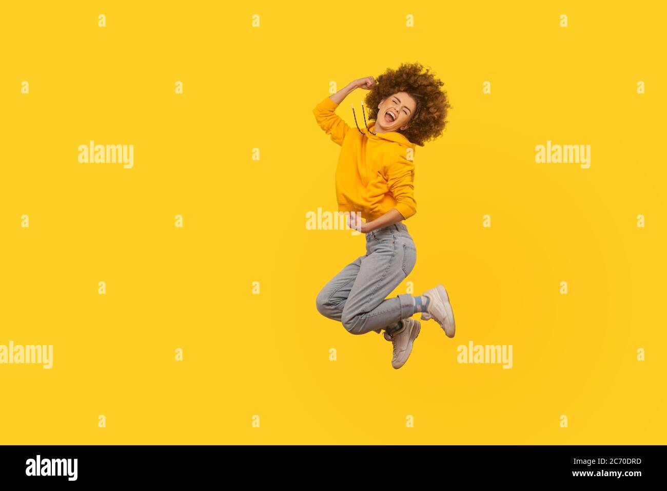 Portrait of overjoyed enthusiastic curly-haired girl in urban style hoodie and jeans jumping high in air, flying and shouting from happiness, rejoicin Stock Photo