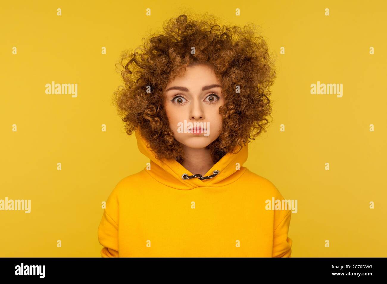 Portrait of cute curly-haired woman in urban style hoodie standing with puffed cheeks, holding breath and looking at camera with big eyes, amazed expr Stock Photo