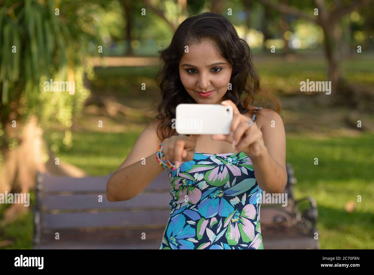 Young beautiful Indian woman taking picture with phone at the park ...
