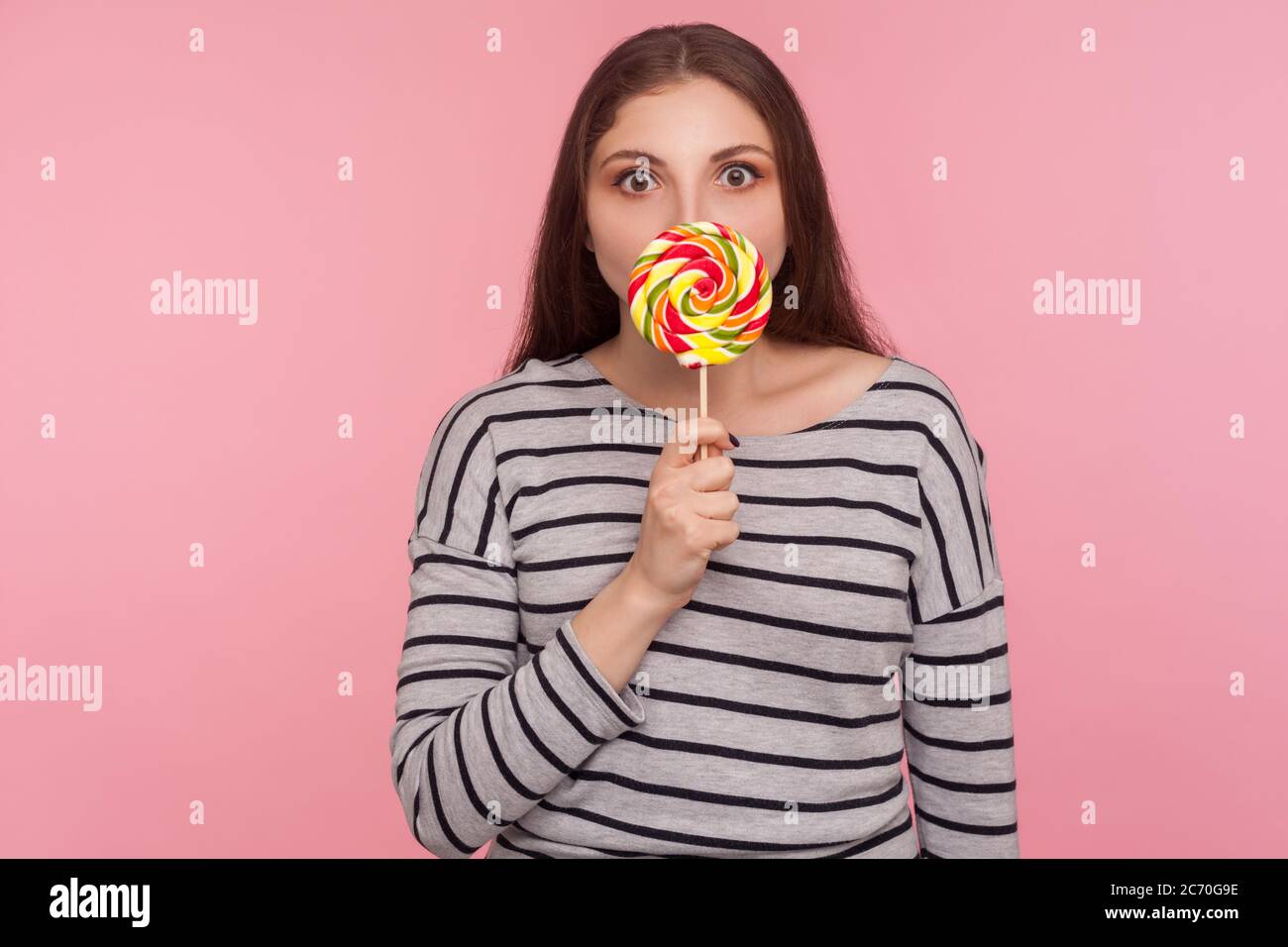 Portrait of amazed woman in striped sweatshirt licking lollipop, tasting sweet round rainbow candy with surprised expression, enjoying delicious flavo Stock Photo