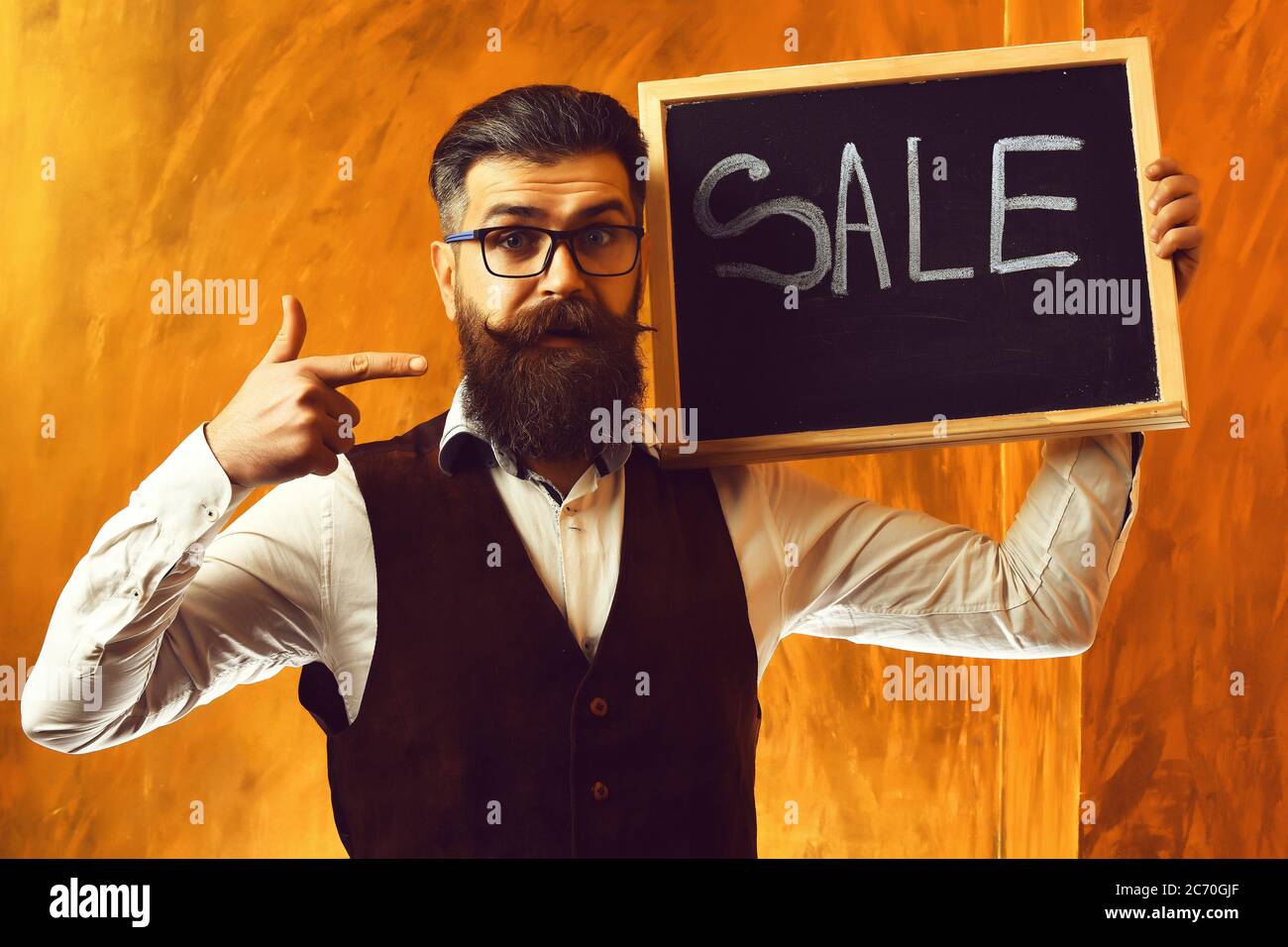 Bearded man, long beard. Brutal caucasian unshaven serious hipster with glasses and moustache holding sale inscription on blackboard wearing white shirt, suede waistcoat on brown studio background Stock Photo