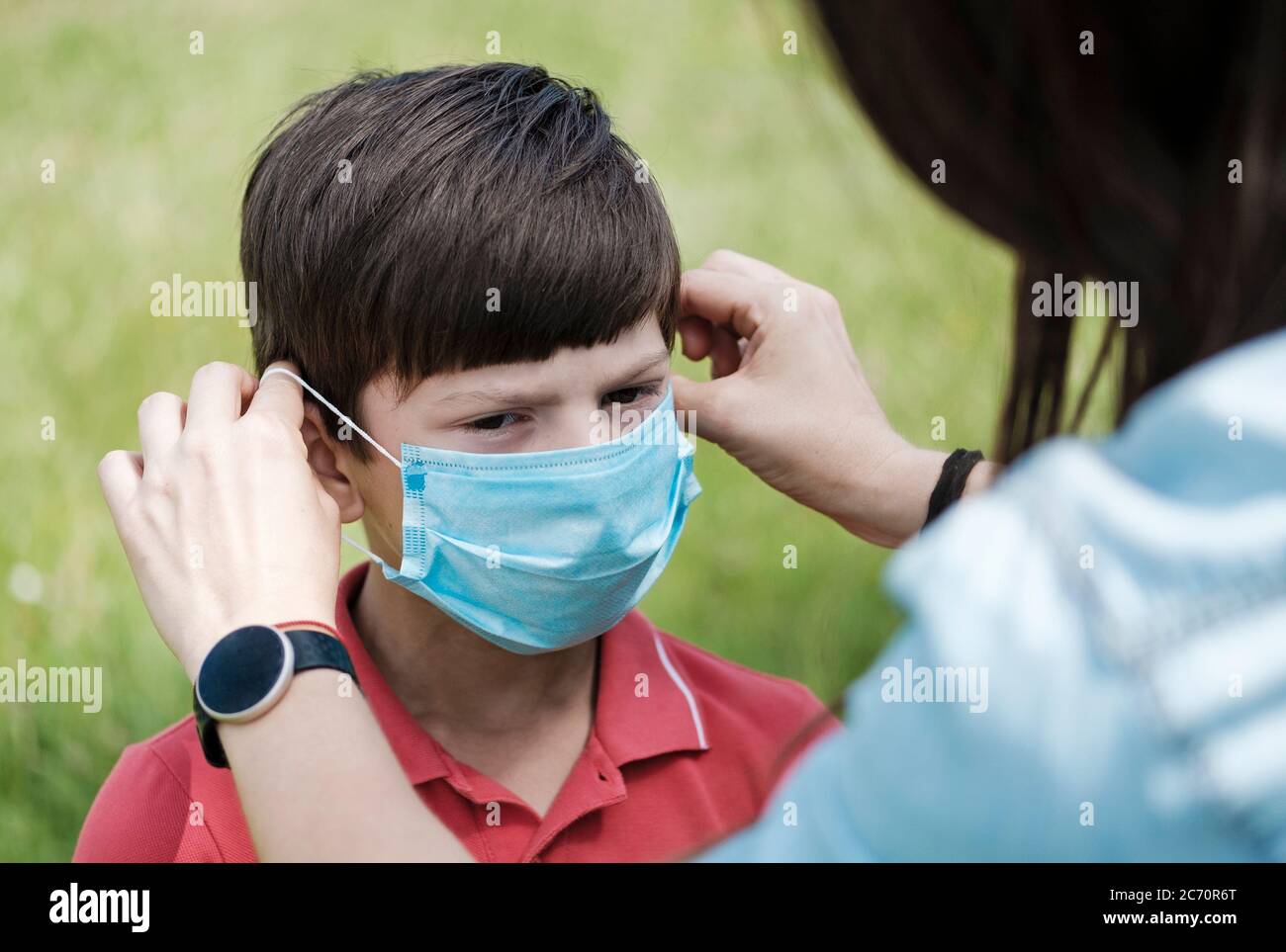 Mother fitting a young boy with a face mask as protection during the Covid-19 or coronavirus pandemic outdoors against green grass in a close up view Stock Photo