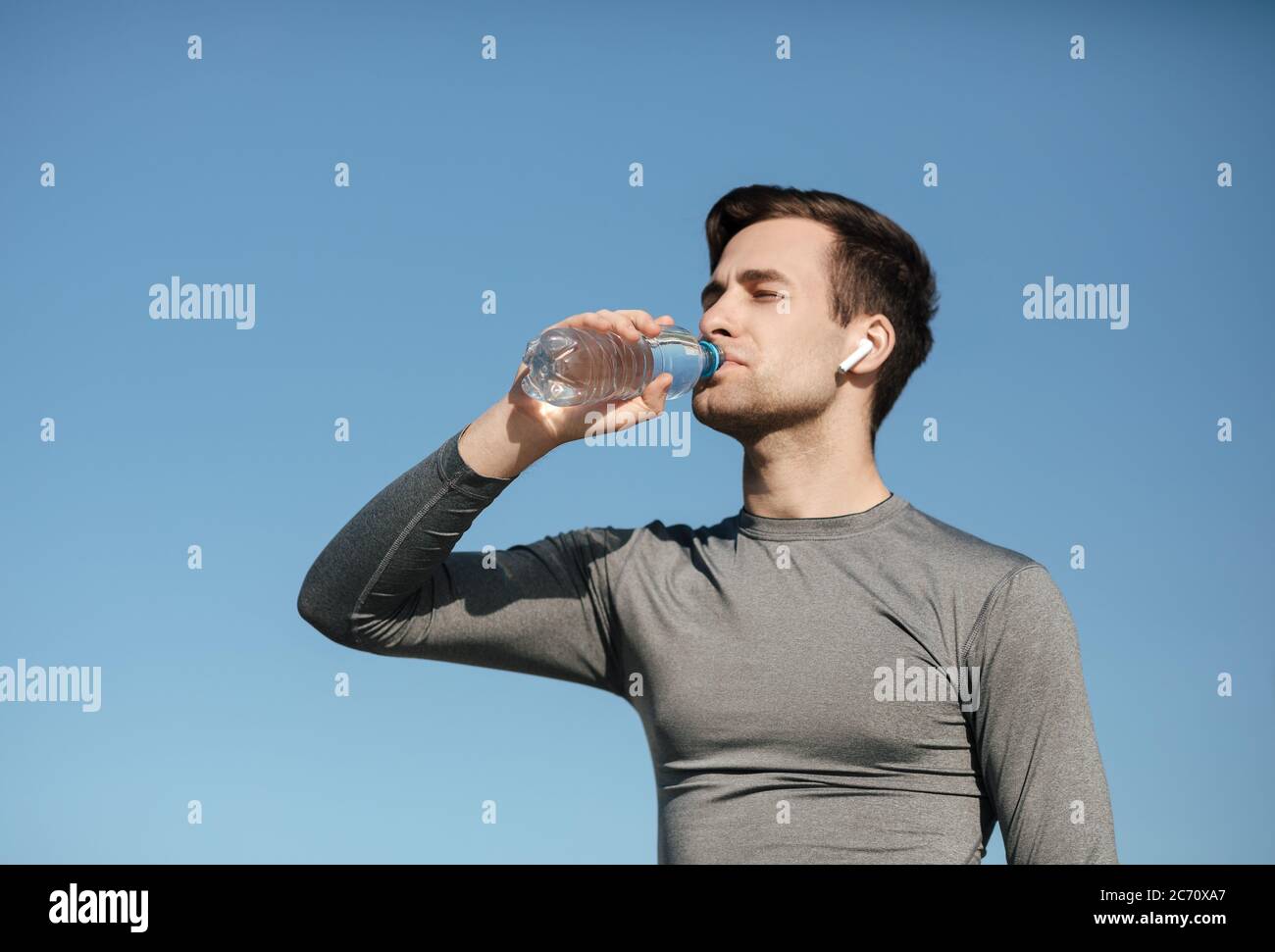 Thirst and sport. Young guy in sport uniform with modern wireless headphones, drinks water Stock Photo