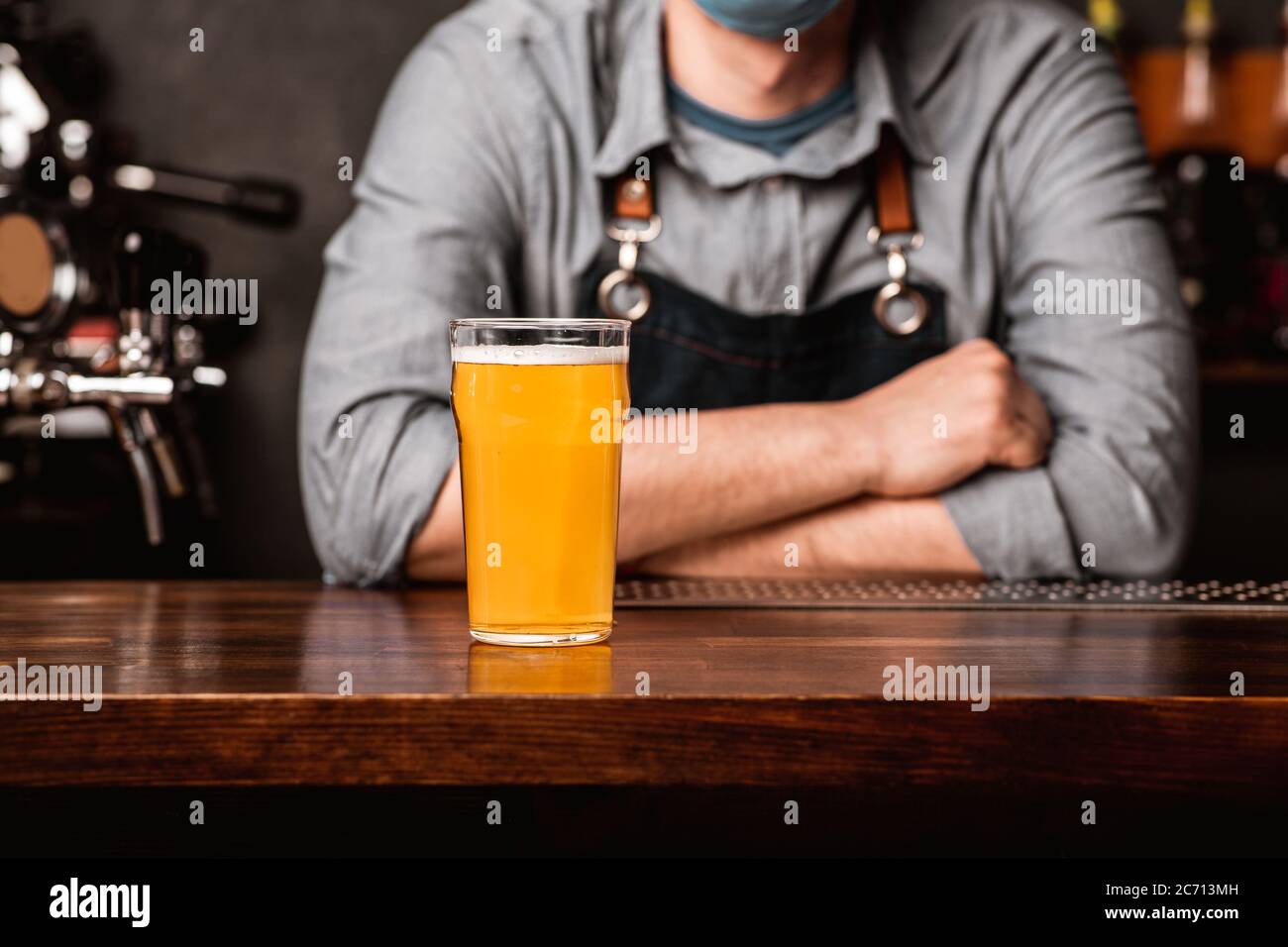 Friendly conversation with bartender. Guy in apron with crossed arms leans on bar and gives glass of lager Stock Photo
