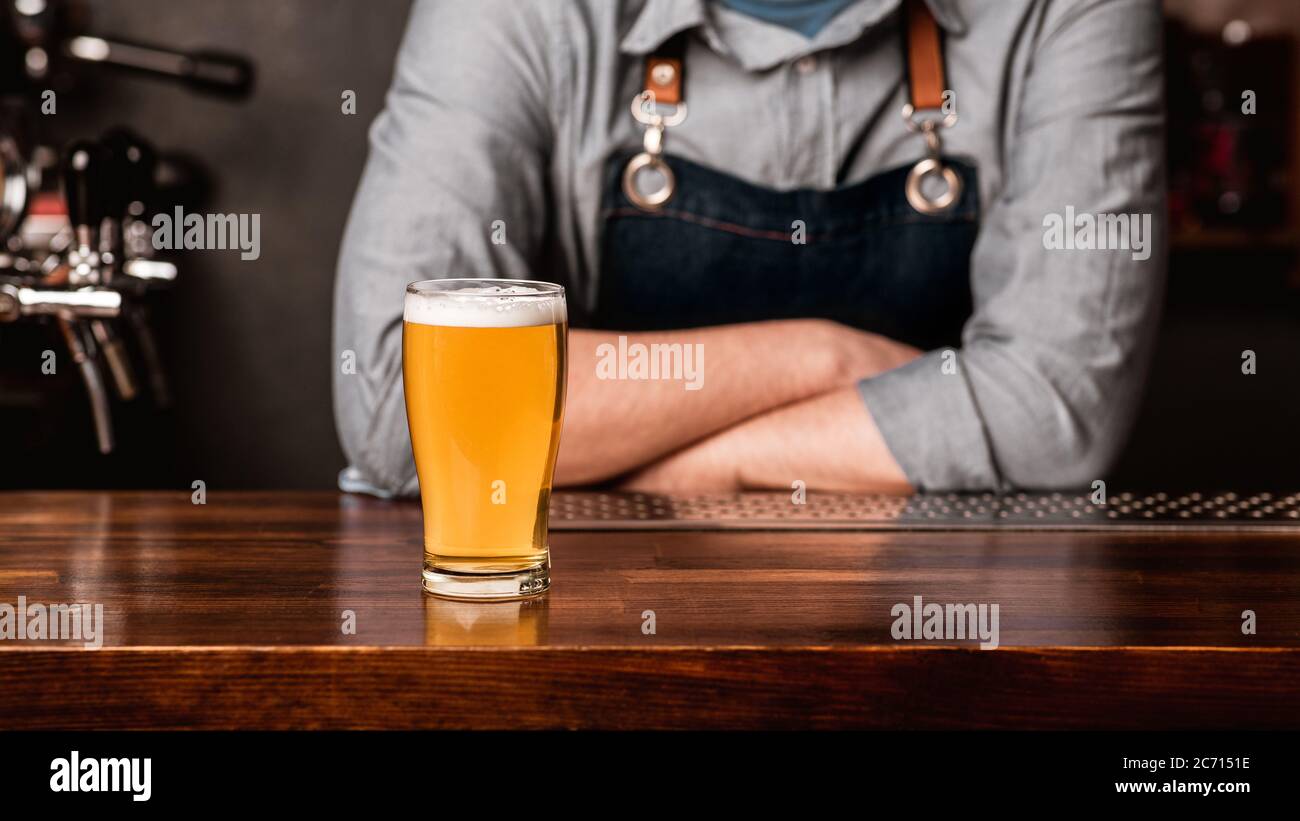Talk with bartender with drink. Man in apron with crossed arms, and light beer with foam on wooden bar counter Stock Photo