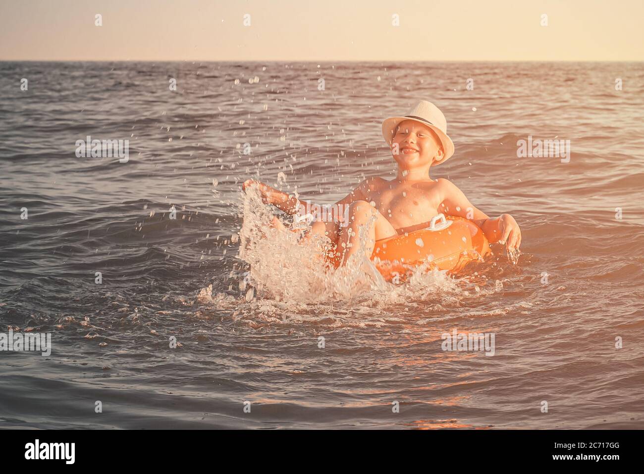 Cheerful Caucasian boy in a white hat on an orange swimming circle splashes his legs in the sea and smile. A child swims and has fun at the resort at Stock Photo