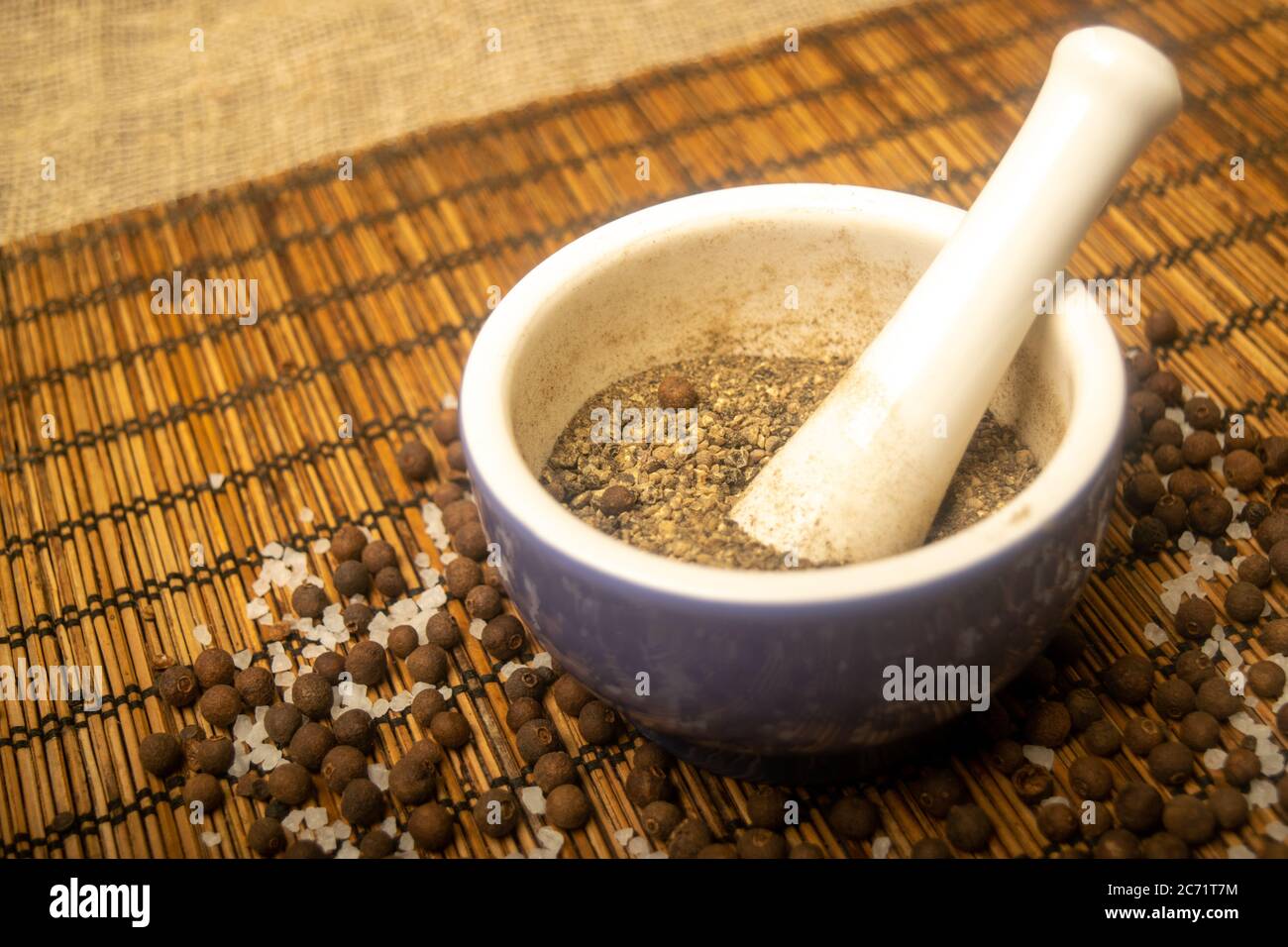 Ground black allspice in a ceramic mortar and black allspice and coarse salt scattered on a reed Mat. Close up Stock Photo