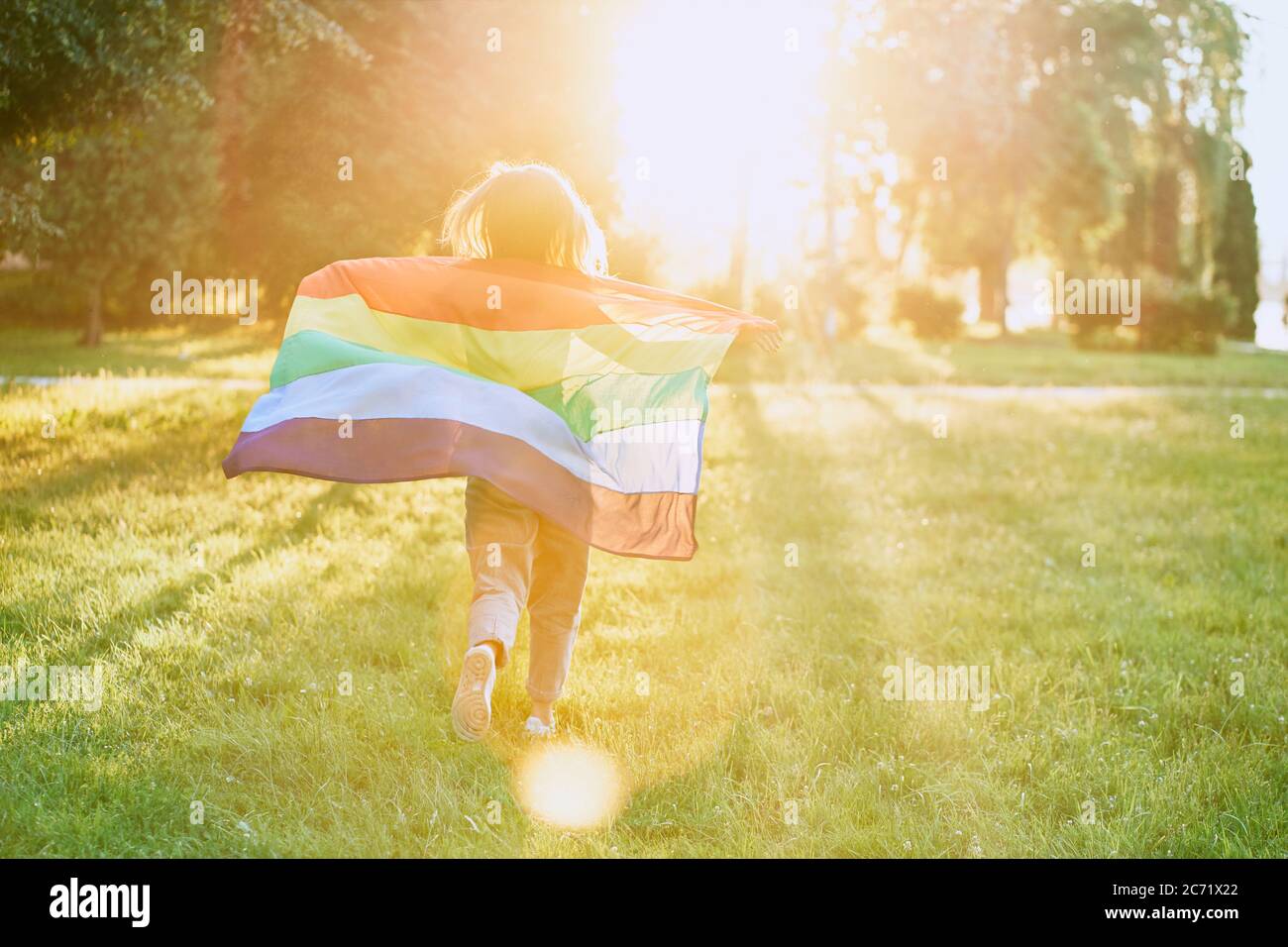 Back view of incognito young woman swinging rainbow flag on wind behind back. Unrecognizable girl running in park, enjoying beautiful summer sunset. Concept of lgbt, minorities tolerance. Stock Photo