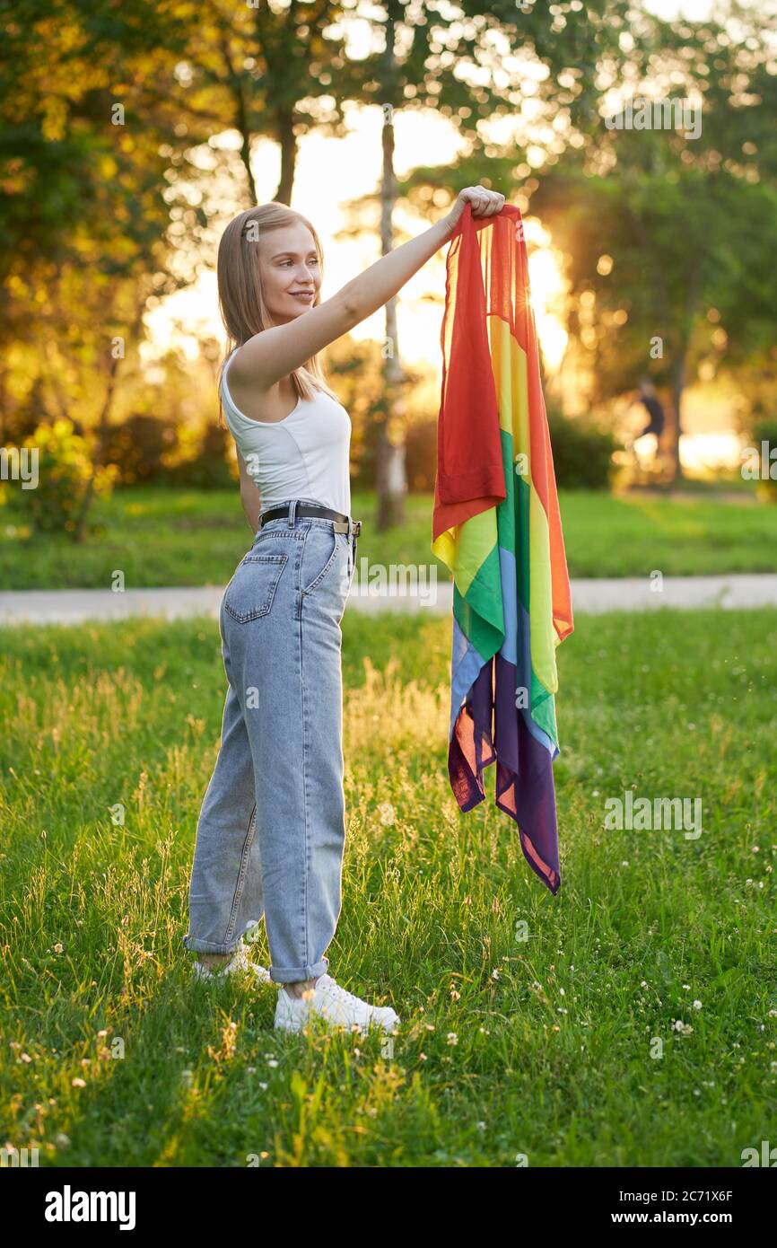 Side view of tolerant girl posing with colorful scarf in summer park, sunset on background. Gorgeous young woman holding rainbow lgbt flag in hand and smiling. Minorities tolerance concept. Stock Photo