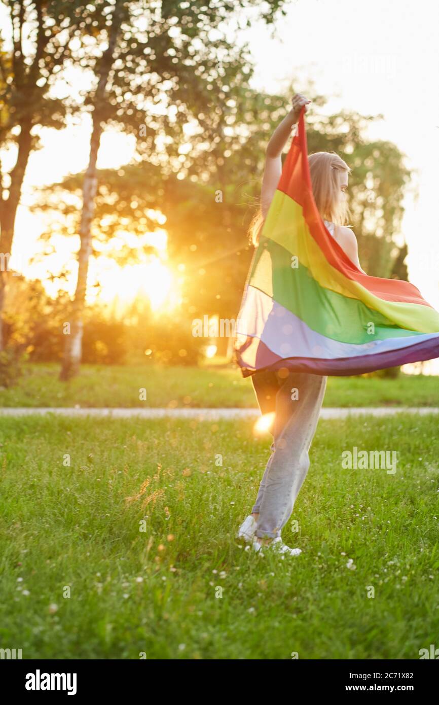 Young unrecogizable woman swinging rainbow lgbt flag on wind behind back while dancing. Back view of girl having fun in park, enjoying summer sunset, full length. Concept of lgbt minorities tolerance. Stock Photo