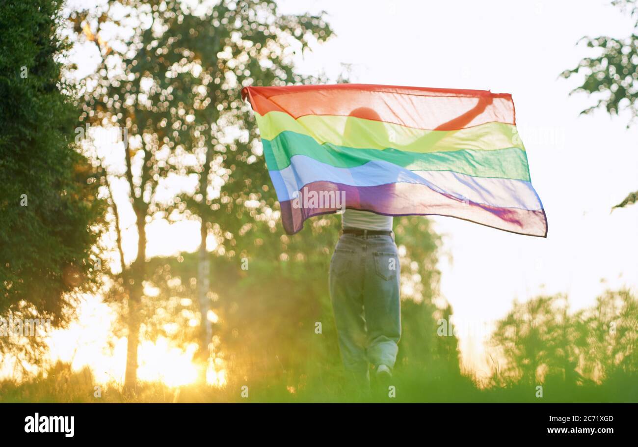 Back view of unrecognizable girl running in park, enjoying summer sunset. Incognito young woman swinging rainbow flag on wind behind back. Concept of lgbt, minorities tolerance. Stock Photo
