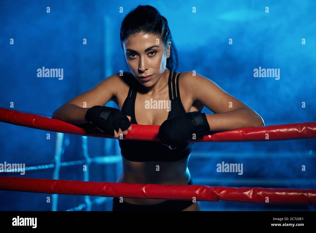 Professional female kickboxer with strong face posing in smoky blue atmosphere, wearing bandages on hands. Fighter having rest after training, leaning on rope and looking at camera. Concept of boxing. Stock Photo