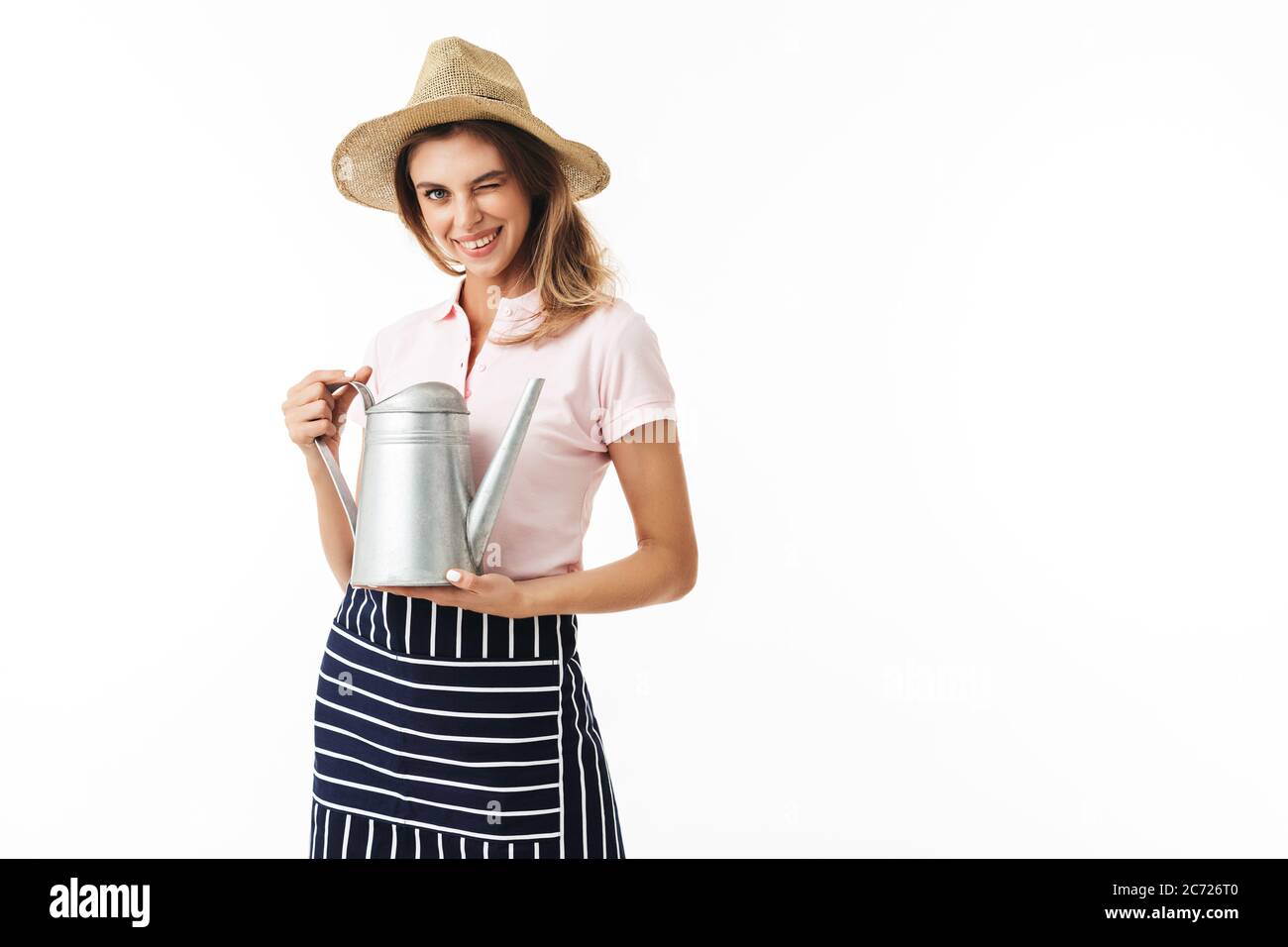 Young smiling woman in straw hat and striped apron happily winking on camera holding watering pot in hands over white background Stock Photo