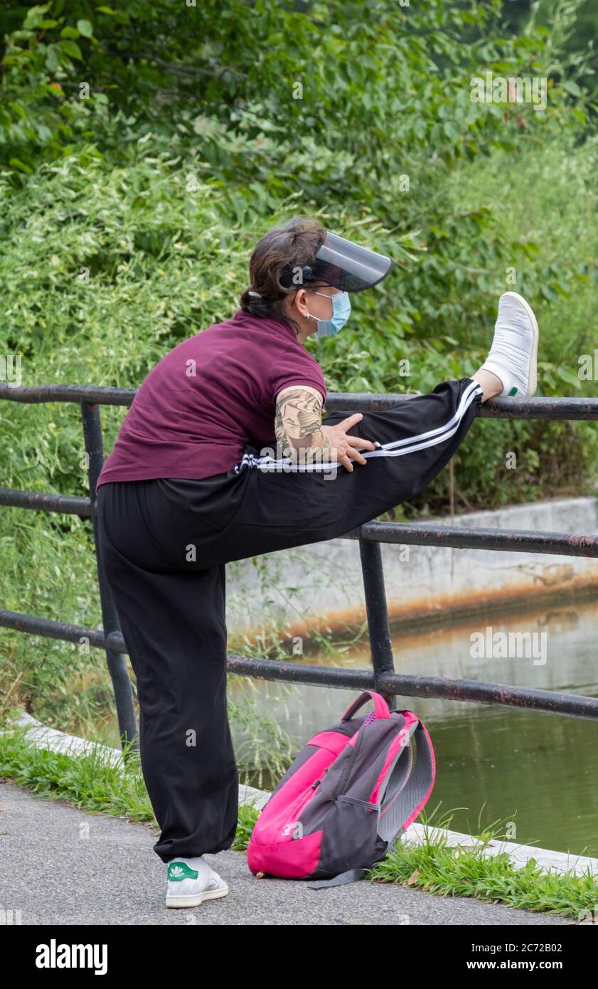 A nimble woman wearing a surgical mask stretches her leg before a morning exercise walk. In Kissena Park, Flushing, Queens, New York Stock Photo