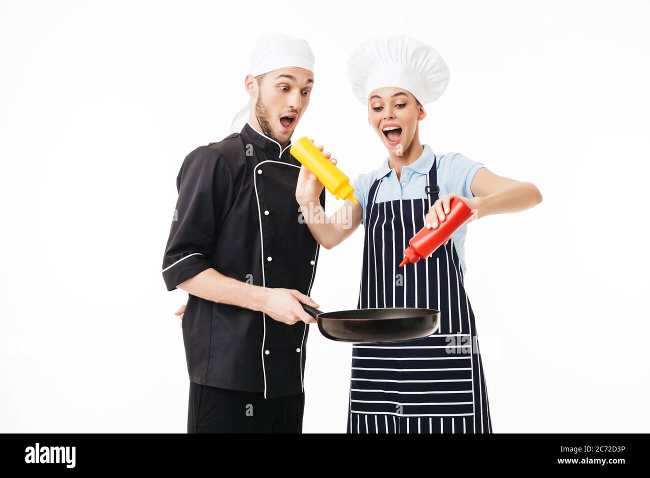 Young shocked man chef in black uniform, holding pan in hands while woman cook in striped apron using bottles of ketchup and mustard on it over white Stock Photo