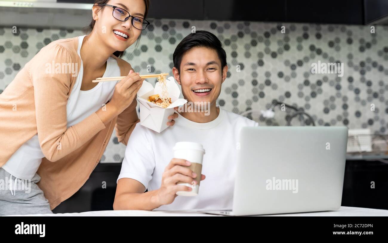 Panoramic asian couple work from home in kitchen and eat delivery chinese take away food and take out coffee while city lockdown from covid-19 pandemi Stock Photo