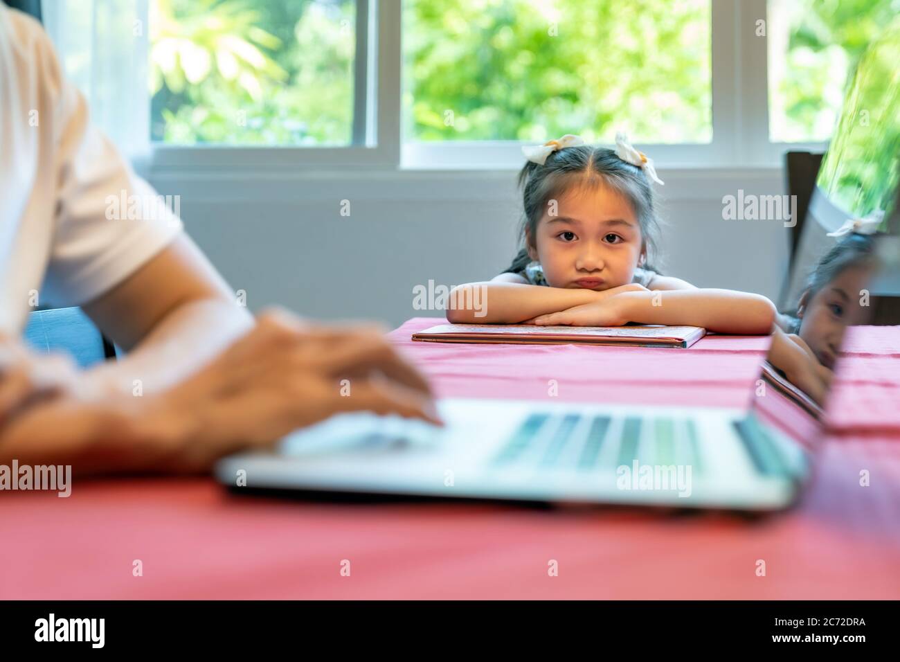 Asian upset girl looking and waiting her father working in dining room. Dad has to working from home while city lockdown from coronavirus covid-19 pan Stock Photo