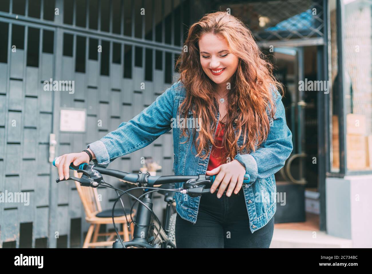 Red curled long hair caucasian teen girl on the city street walking with bicycle fashion portrait. Natural people beauty urban life concept image. Stock Photo