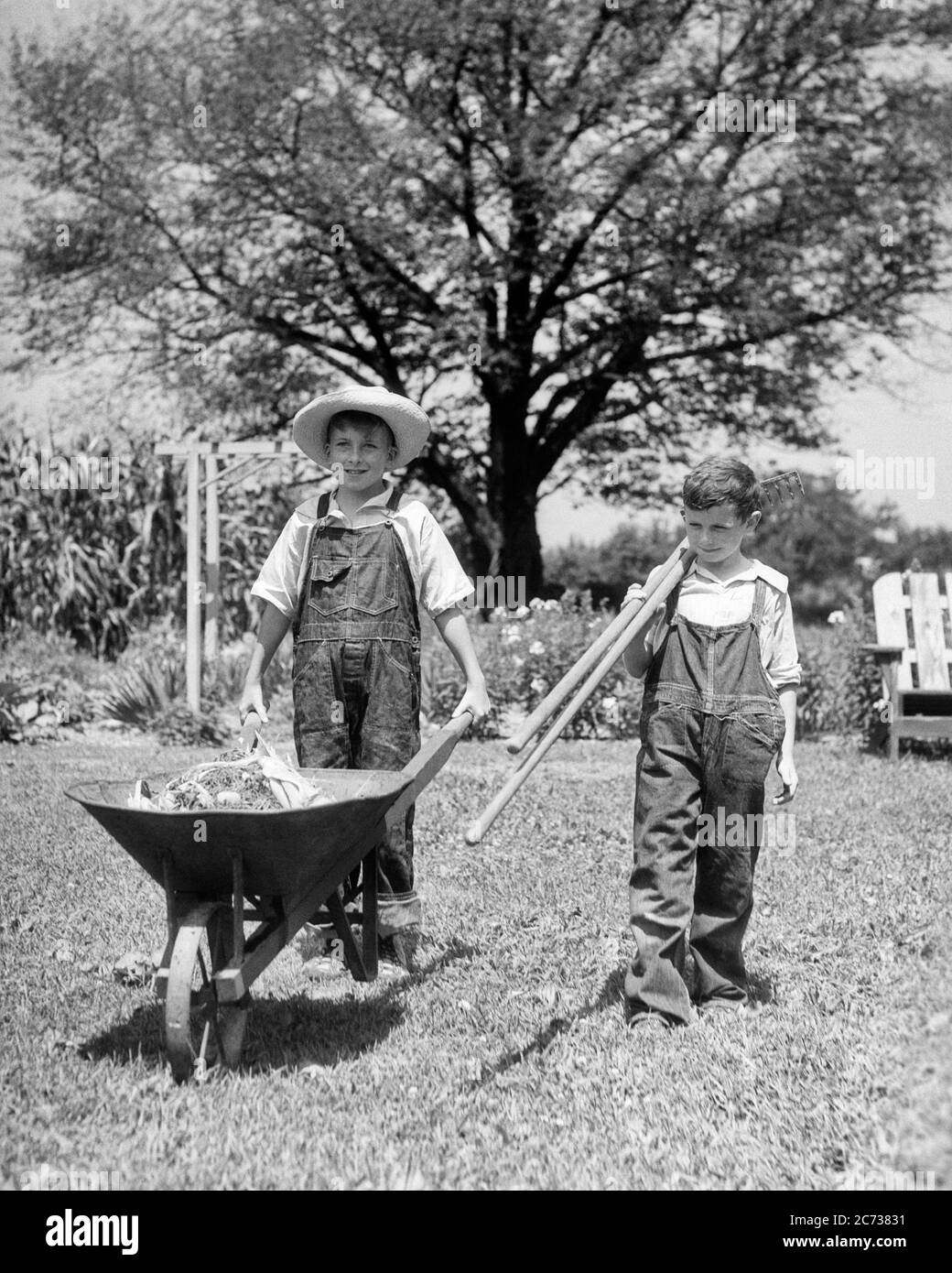 1930s TWO YOUNG SMILING FARM BOYS BROTHERS WEARING OVERALLS WALKING TOGETHER TOWARD CAMERA WITH WHEELBARROW AND GARDENING TOOLS  - f6904 HAR001 HARS PLEASED JOY LIFESTYLE BROTHERS JOBS RURAL HEALTHINESS HOME LIFE COPY SPACE FRIENDSHIP FULL-LENGTH OVERALLS CARING FARMING MALES SIBLINGS WHEELBARROW CONFIDENCE AGRICULTURE B&W GOALS HAPPINESS WELLNESS CHEERFUL STRENGTH STRATEGY AND FARMERS LEADERSHIP TOWARD PRIDE OPPORTUNITY SIBLING SMILES TASKS CONNECTION CONCEPTUAL STRAW HAT JOYFUL STYLISH VARIOUS BLUE JEANS COOPERATION GROWTH JUVENILES TOGETHERNESS BLACK AND WHITE CAUCASIAN ETHNICITY HAR001 Stock Photo