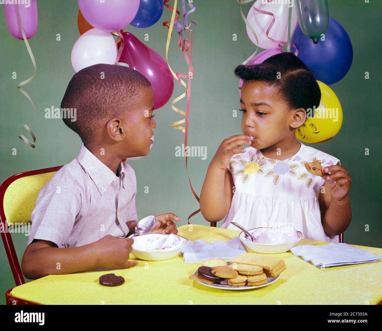 1960s AFRICAN-AMERICAN BOY AND GIRL SHARING EATING ICE CREAM AND COOKIES TOGETHER AT BIRTHDAY PARTY  - kn530 HAR001 HARS STREAMERS STUDIO SHOT HOME LIFE COPY SPACE FRIENDSHIP MALES COOKIES SIBLINGS SISTERS HEAD AND SHOULDERS HIGH ANGLE AFRICAN-AMERICANS AFRICAN-AMERICAN AND EXCITEMENT BLACK ETHNICITY PARTIES SIBLING CONNECTION FRIENDLY ICE CREAM GROWTH JUVENILES TOGETHERNESS HAR001 OLD FASHIONED AFRICAN AMERICANS Stock Photo
