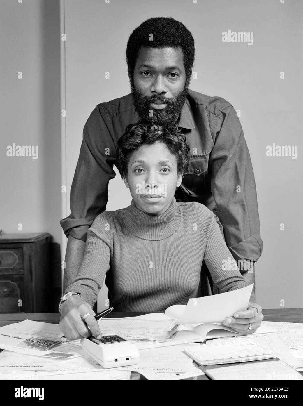 1970s AFRICAN-AMERICAN COUPLE LOOKING AT CAMERA MAN STANDING BEHIND WOMAN SITTING AT TABLE WITH PAPERS BILLS HAND ON CALCULATOR - s21331 HAR001 HARS CALCULATOR FACIAL BUDGET STYLE COMMUNICATION TEAMWORK LIFESTYLE FEMALES MARRIED SPOUSE HUSBANDS HOME LIFE COPY SPACE FRIENDSHIP HALF-LENGTH LADIES PERSONS MALES RISK EXPRESSIONS B&W PARTNER EYE CONTACT GOALS AFRICAN-AMERICANS AFRICAN-AMERICAN BLACK ETHNICITY INNOVATION FACIAL HAIR CONNECTION BEARDS COOPERATION MID-ADULT MID-ADULT MAN MID-ADULT WOMAN SOLUTIONS TOGETHERNESS WIVES BLACK AND WHITE HAR001 OLD FASHIONED TURTLENECK UNSMILING Stock Photo