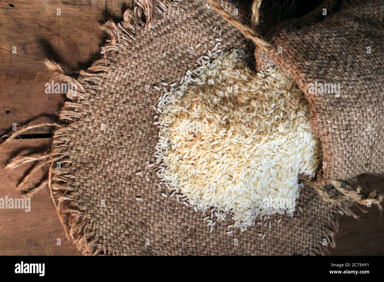 Rice out from small sack isolated on wooden background. Fresh rice in sack bag over wooden textured background. Close up raw rice grain on sack cloth. Stock Photo