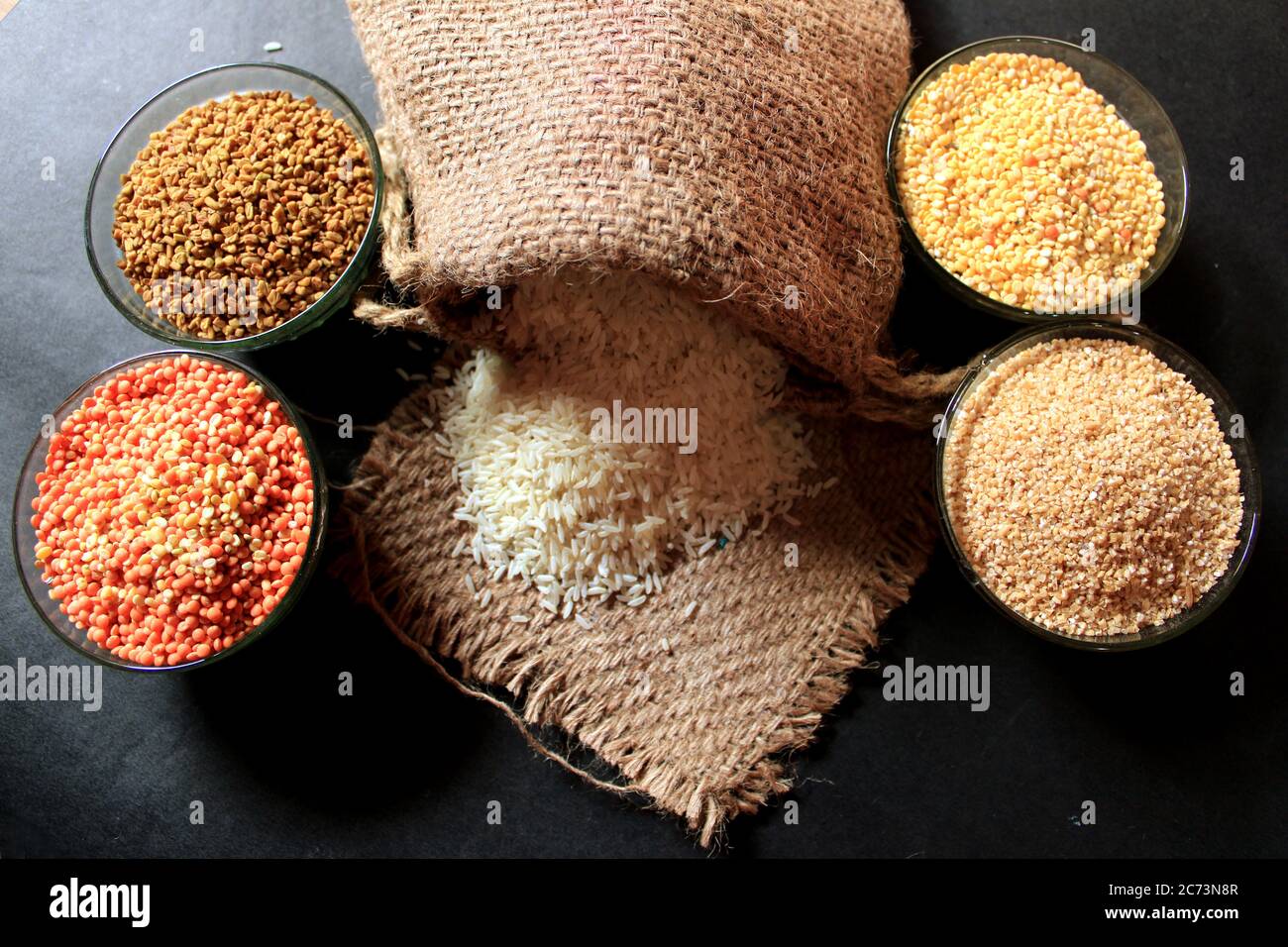 Rice out from small sack isolated on black background. Different colorful pulse in a bowl. Lentil, Fenugreek Seed, Wheat Dalia, Pigeon pea Stock Photo