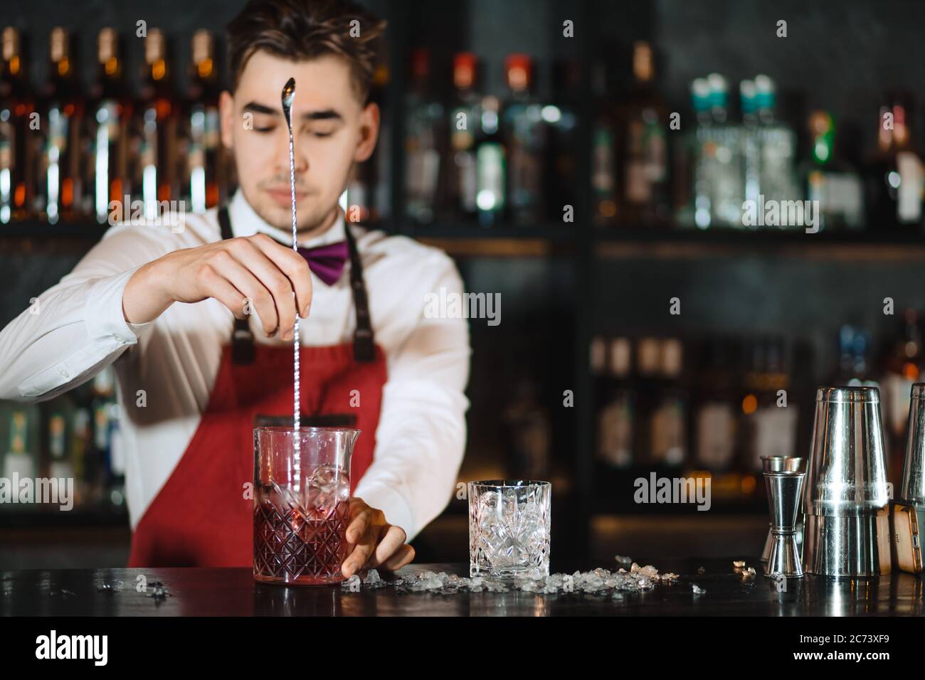 Young professional bartender mixing with long metal spoon cocktail in a glass standing at wooden