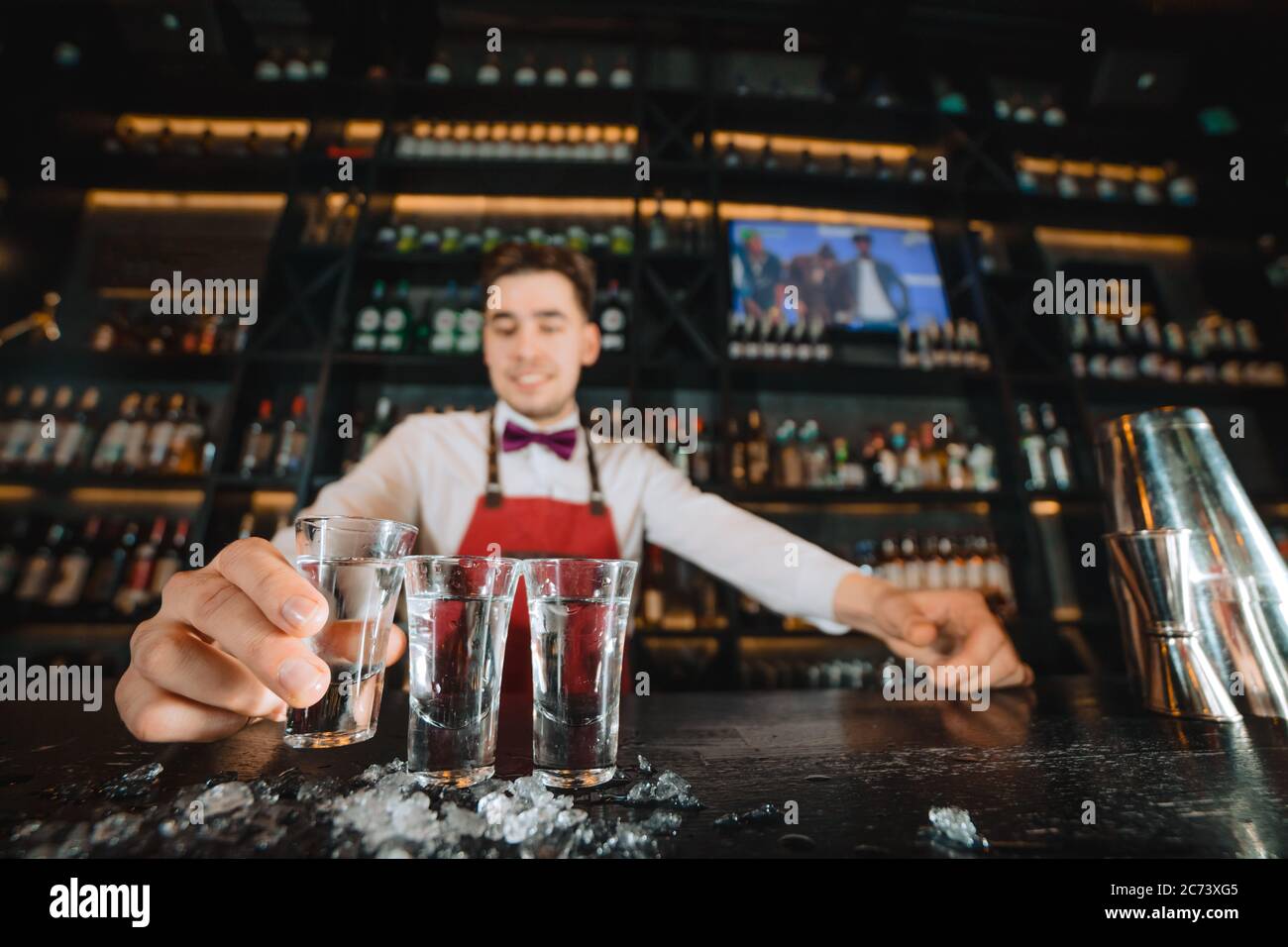 Close-up of expert bartender working in a bar. Focus on shots with vodka or another strong alcohol, served by smiling pleasant barkeeper. Stock Photo