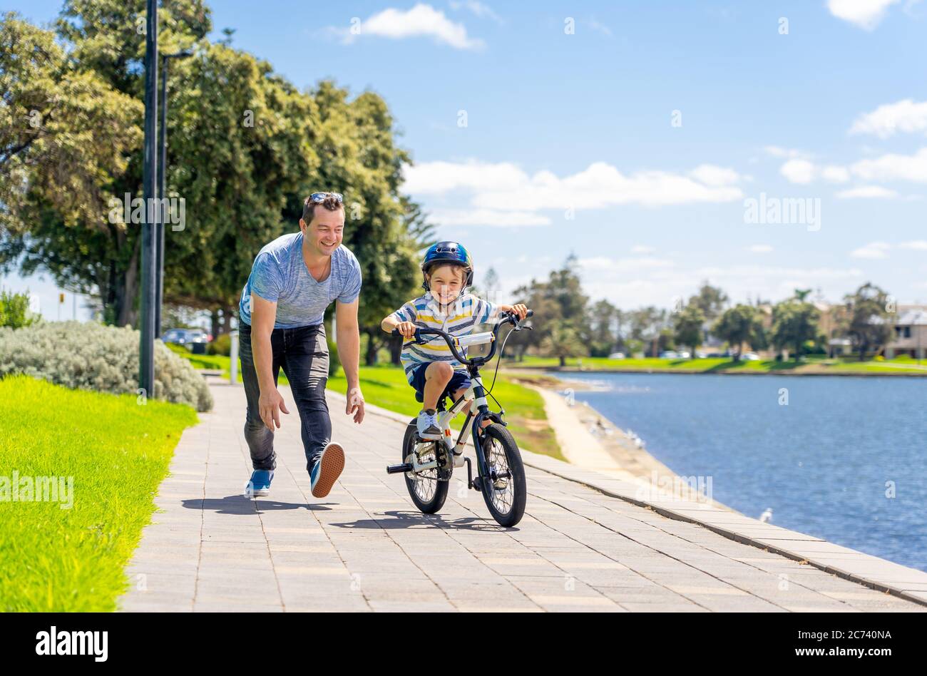 Boy learning to ride a bicycle with his father in the park by the lake. Father and son having fun together on the bikes. Happy family, outdoors activi Stock Photo