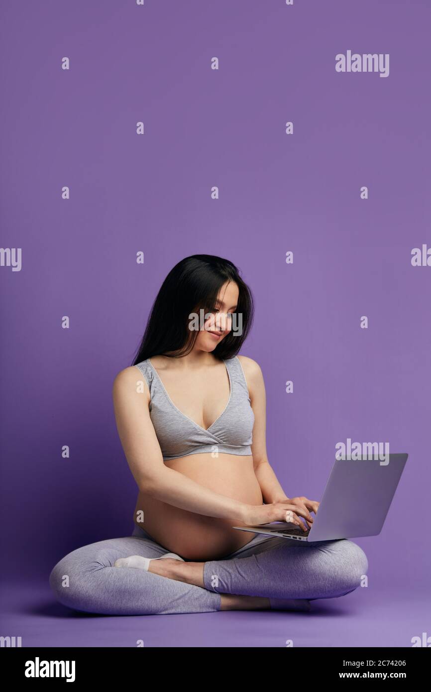 Pregnant young Asian woman in yoga outfit sitting in lotus pose and  browsing internet on laptop in purple studio background Stock Photo - Alamy