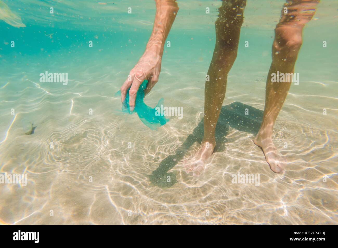 Man collects packages from the beautiful turquoise sea. Paradise beach pollution. Problem of spilled rubbish trash garbage on the beach sand caused by Stock Photo