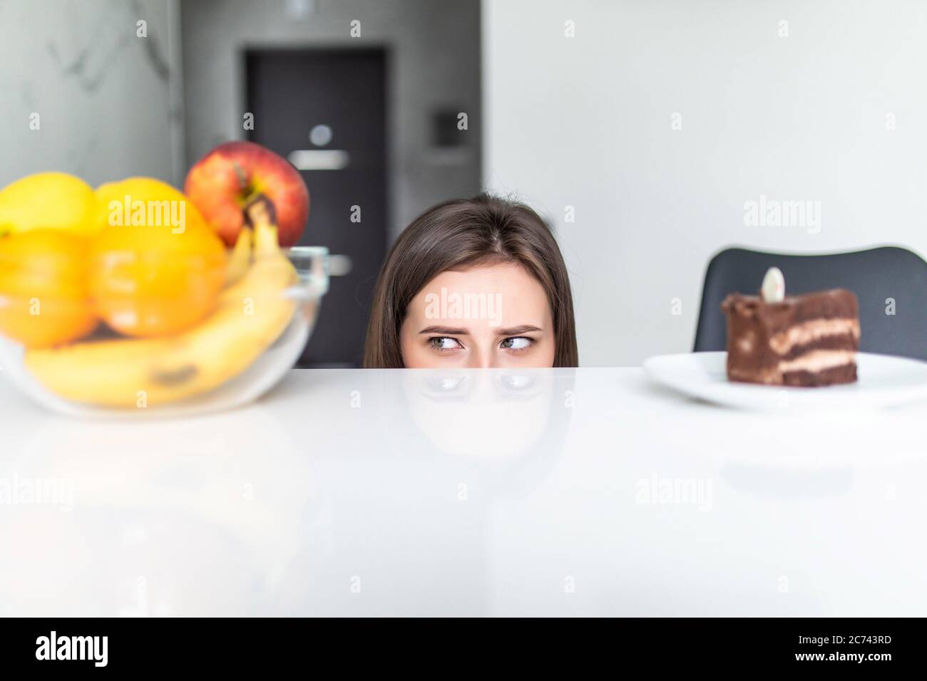 Hard choice. Yound sporty woman is choosing between fruits and sweets while standing on light kitchen. Stock Photo