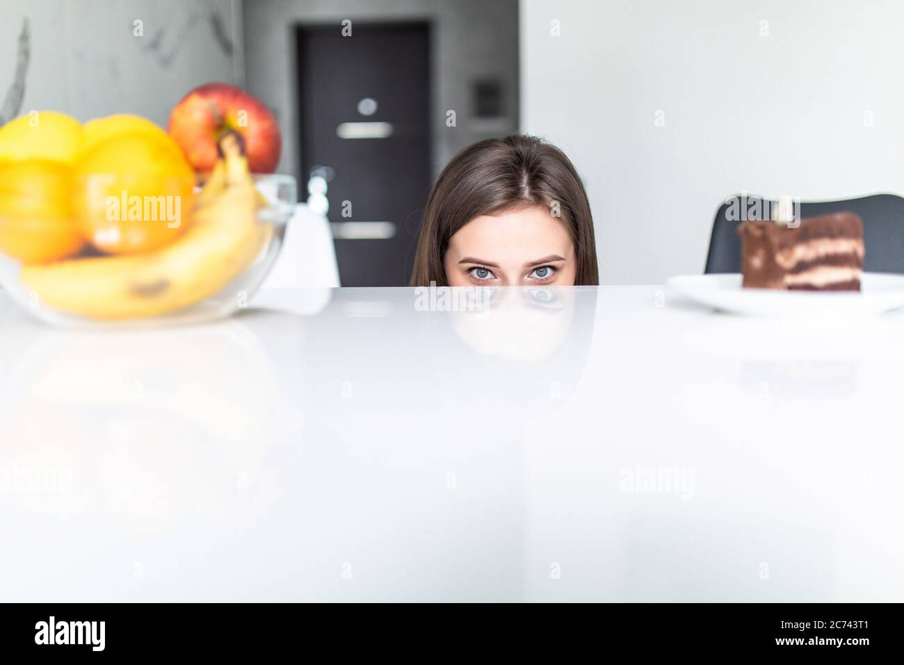 Hard choice. Yound sporty woman is choosing between fruits and sweets while standing on light kitchen. Stock Photo