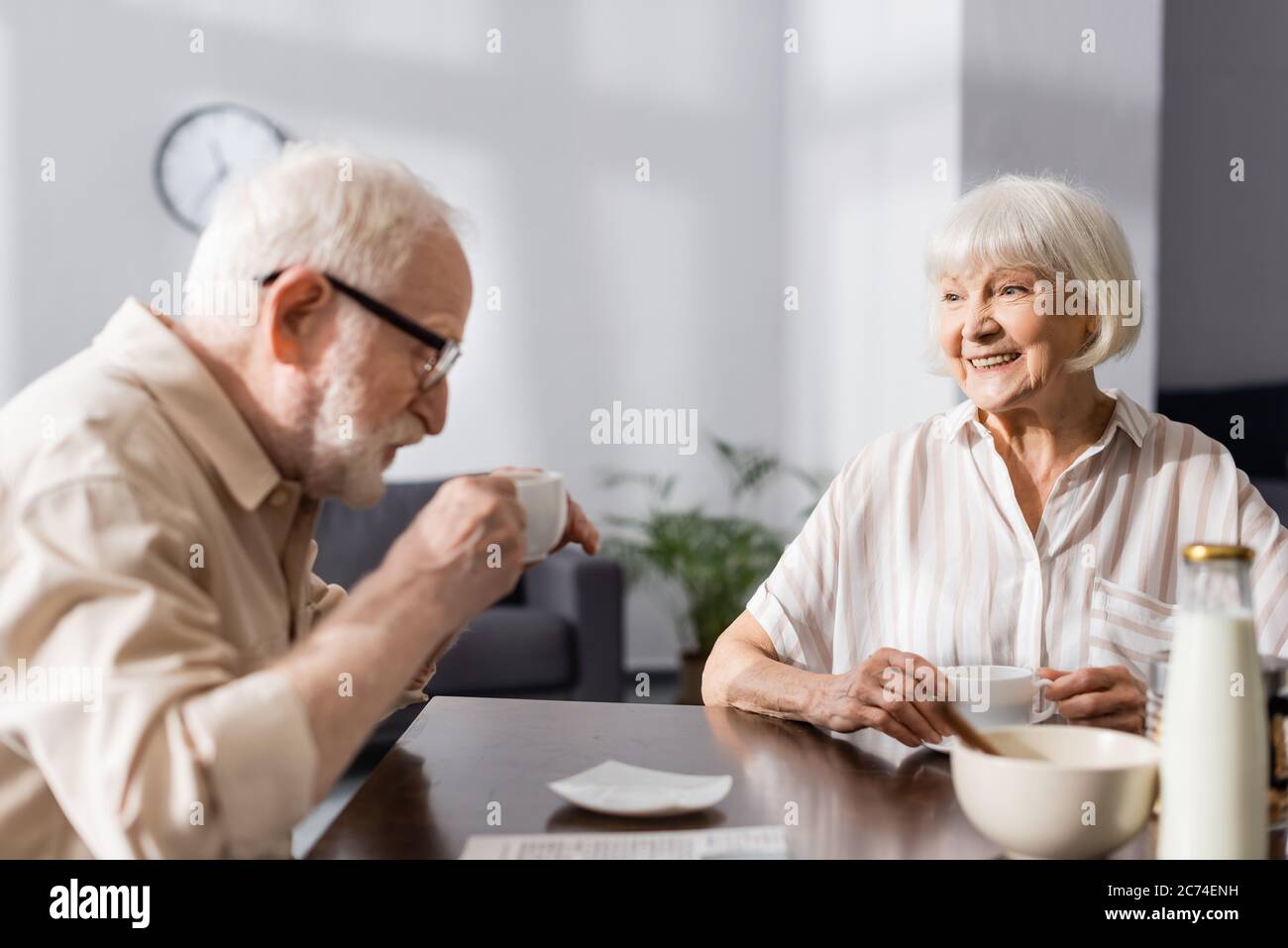 Selective focus of smiling senior woman looking at husband drinking coffee at home Stock Photo
