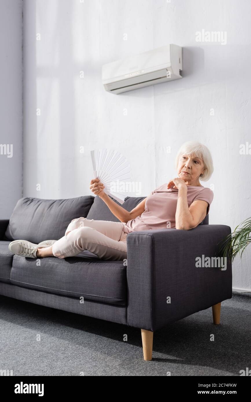 Elderly woman waving fan under air conditioner in living room Stock