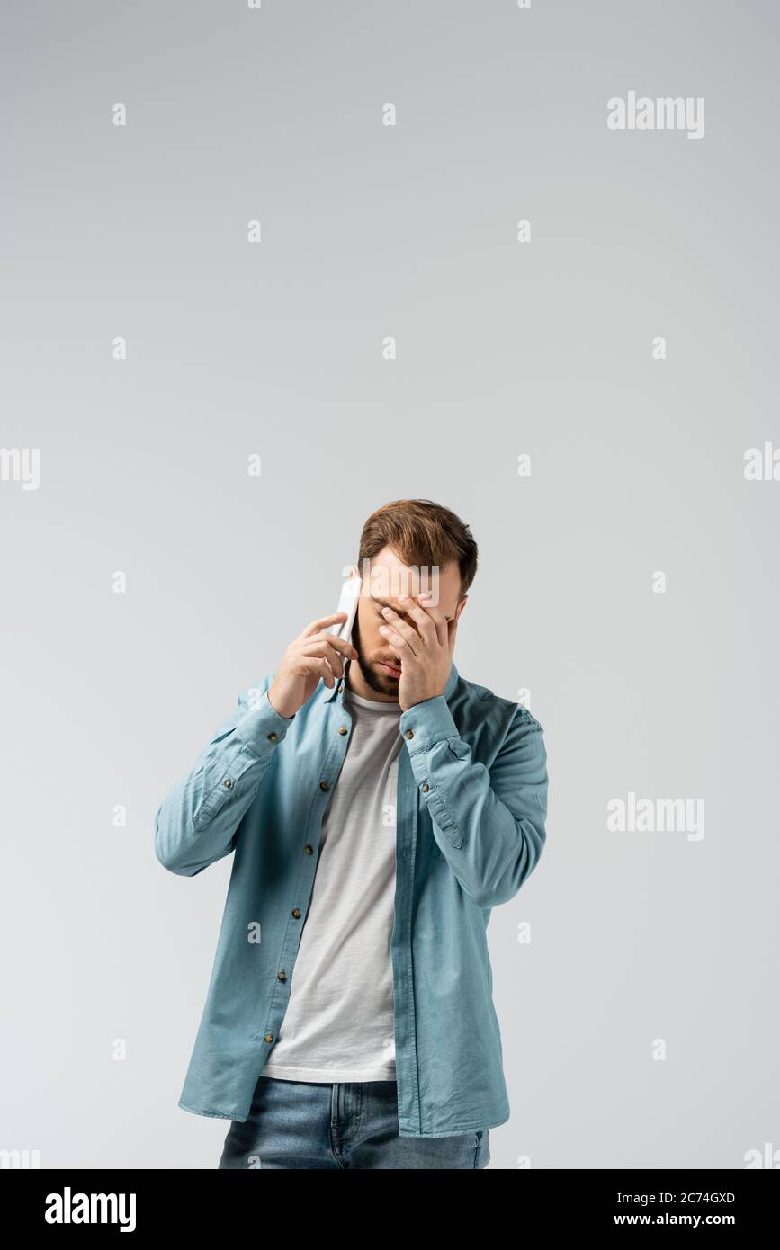 confused young man talking on smartphone and showing facepalm isolated on grey Stock Photo