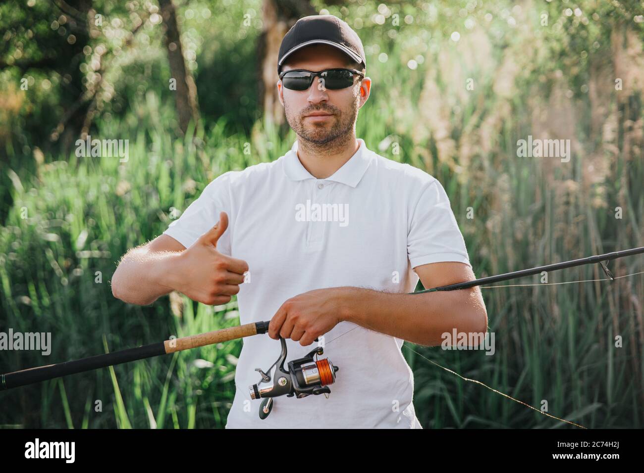 https://c8.alamy.com/comp/2C74H2J/young-fisherman-fishing-on-lake-or-river-guy-stand-alone-and-hold-big-thumb-up-carry-long-rod-with-some-reel-underneath-posing-on-camera-before-cat-2C74H2J.jpg