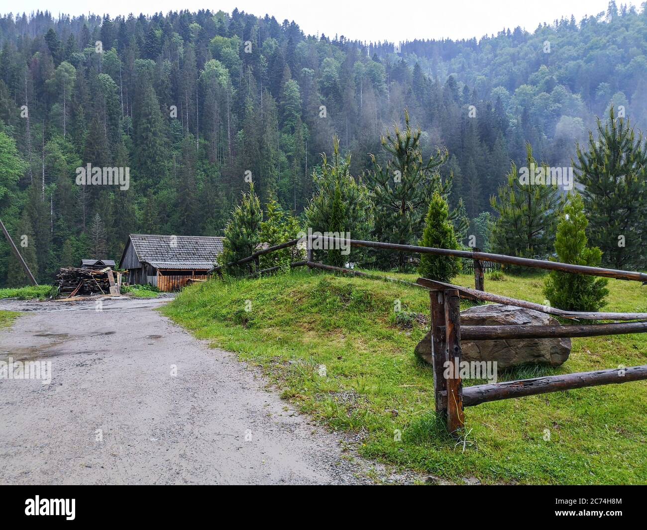 Road along rustic log fence running into a hut and stack of wood amidst evergreen forest at the bottom of Carpathian mountains. Lonely quiet retreat. Stock Photo