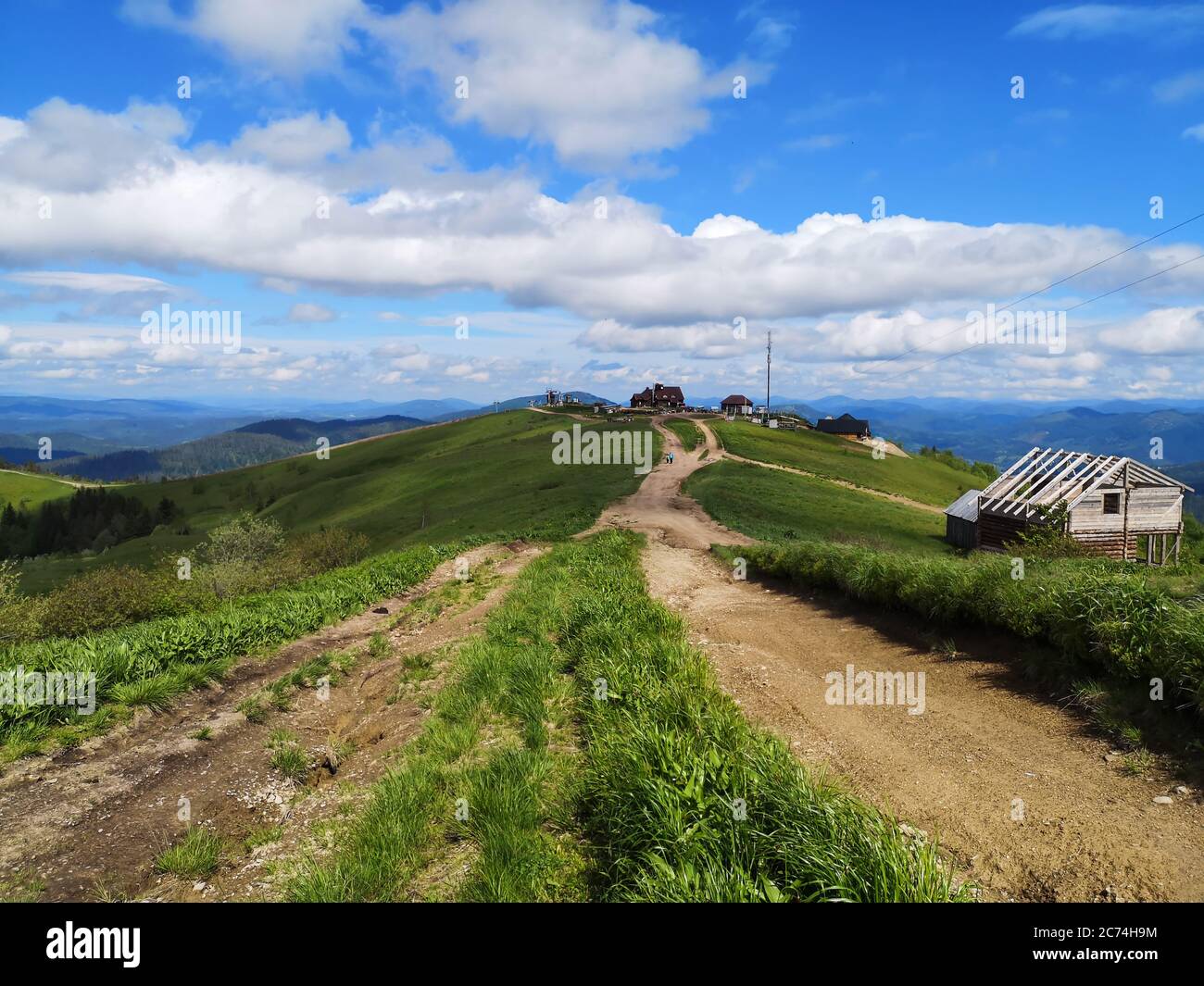 Earthy empty road along the top of Carpathian mountains high below white puffy clouds. Timber wooden houses on the horizon and on the roadside. Stock Photo