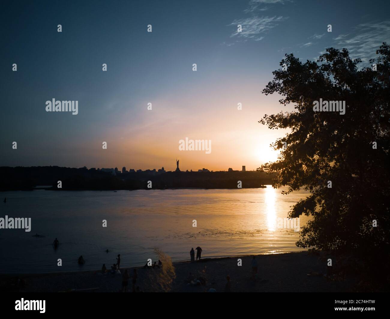 People swimming and having rest on the beach on the left bank of the Dnipro river in Kyiv on summer evening. Urban cityscape with motherland monument. Stock Photo