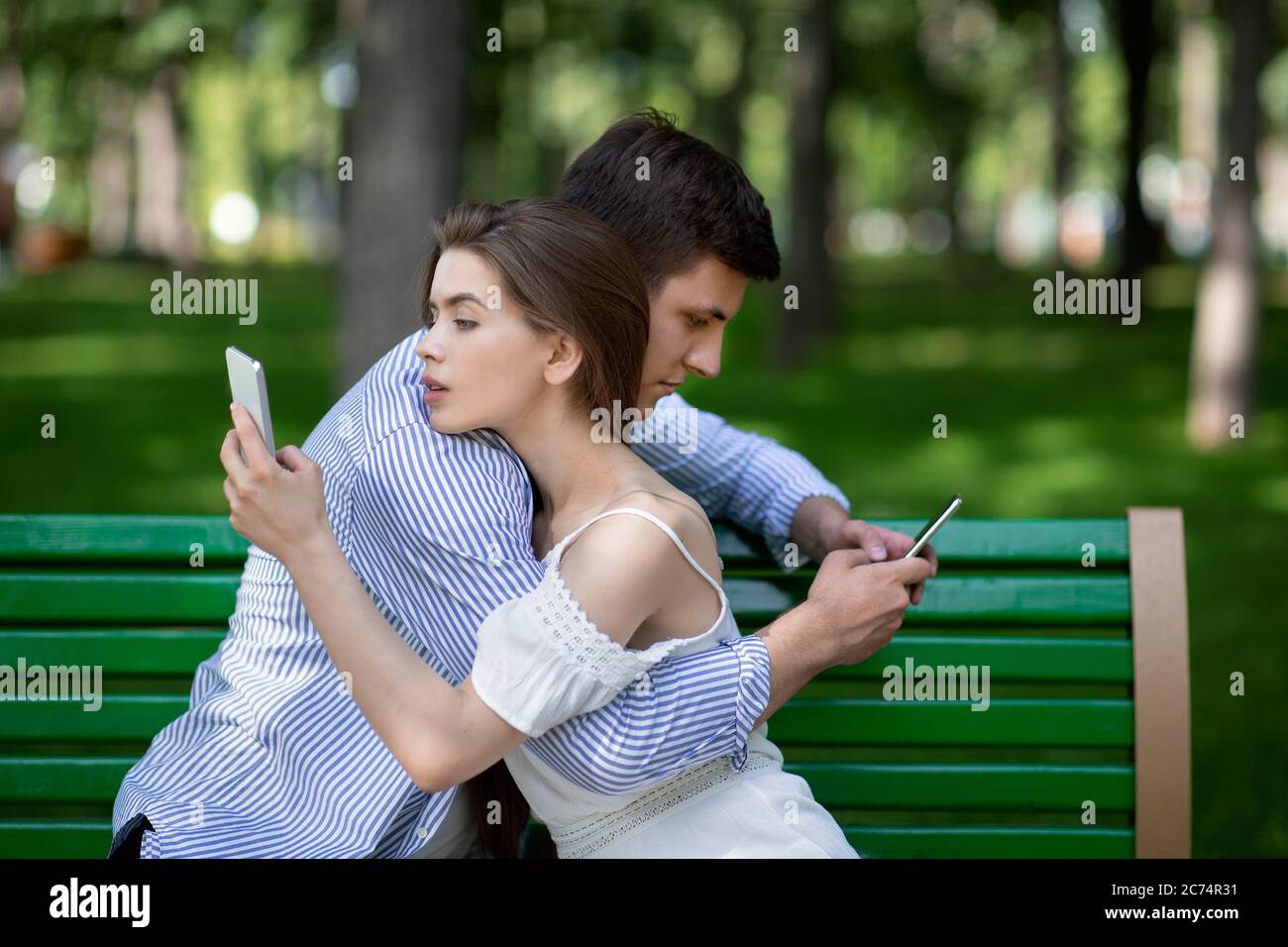 Gadget addicted couple using mobile phones while embracing on bench at park Stock Photo