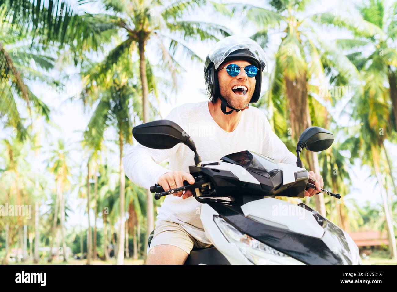 Happy smiling and screaming male tourist in helmet and sunglasses riding motorbike  scooter during his tropical vacation under palm trees Stock Photo - Alamy