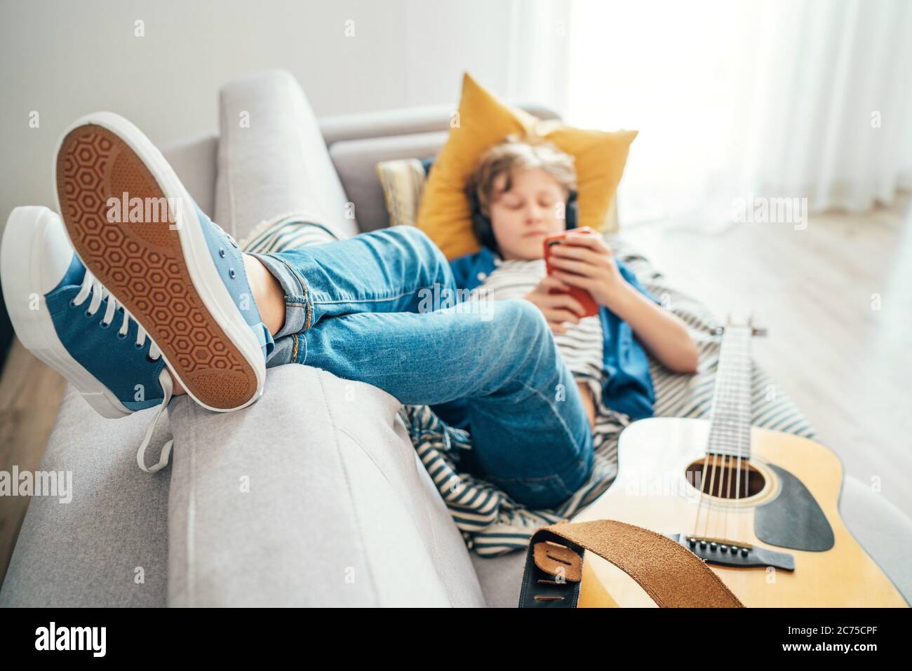 Preteen boy lying with guitar on cozy sofa dressed casual jeans and new sneakers listening to music and chatting using wireless headphones connected w Stock Photo