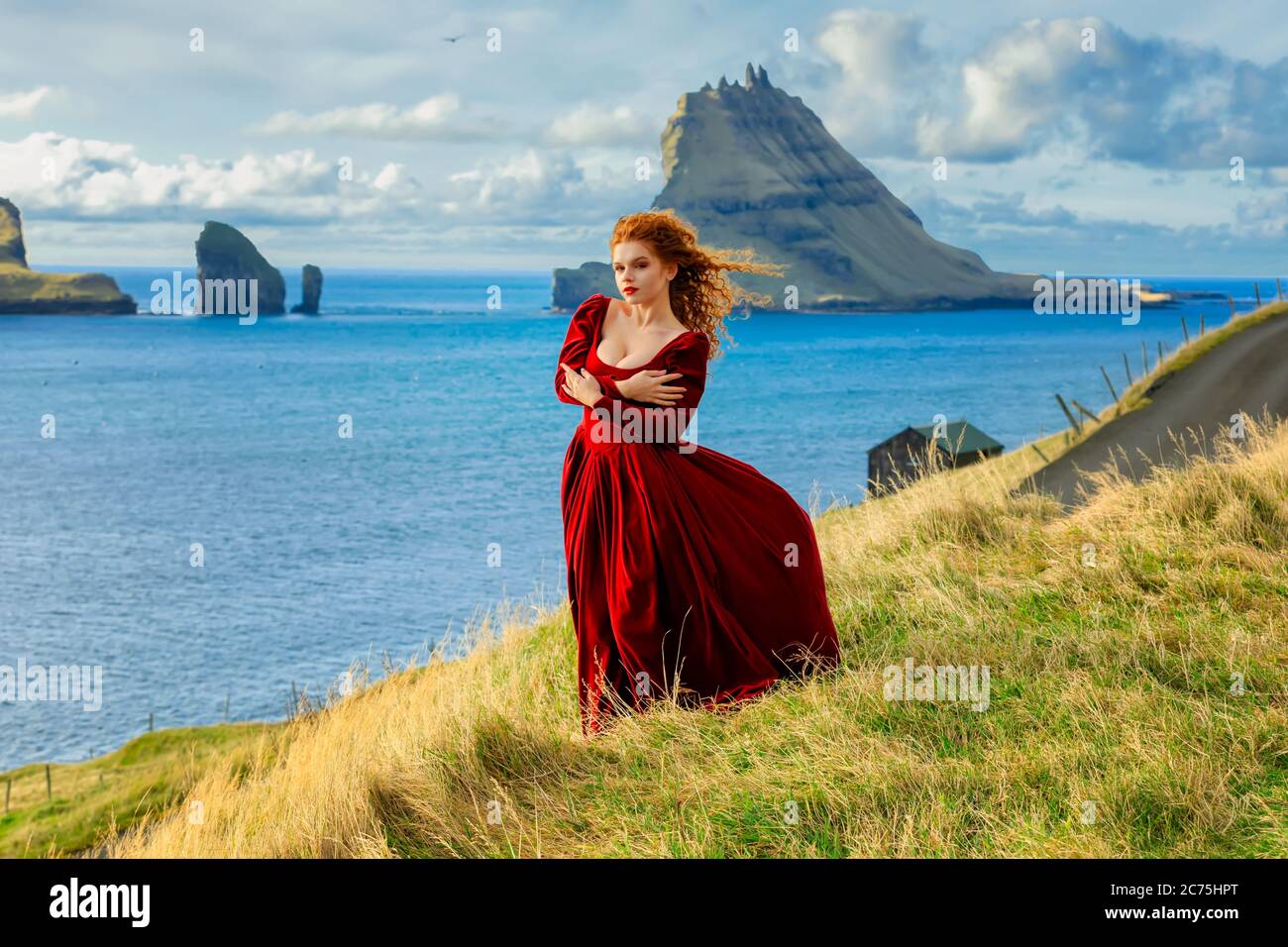 A young redhead woman stays on cliff near Tindholmur island in old-fashioned clothes. Atlantic ocean coastline, Island Vagar, Faroe Islands. Europe Stock Photo