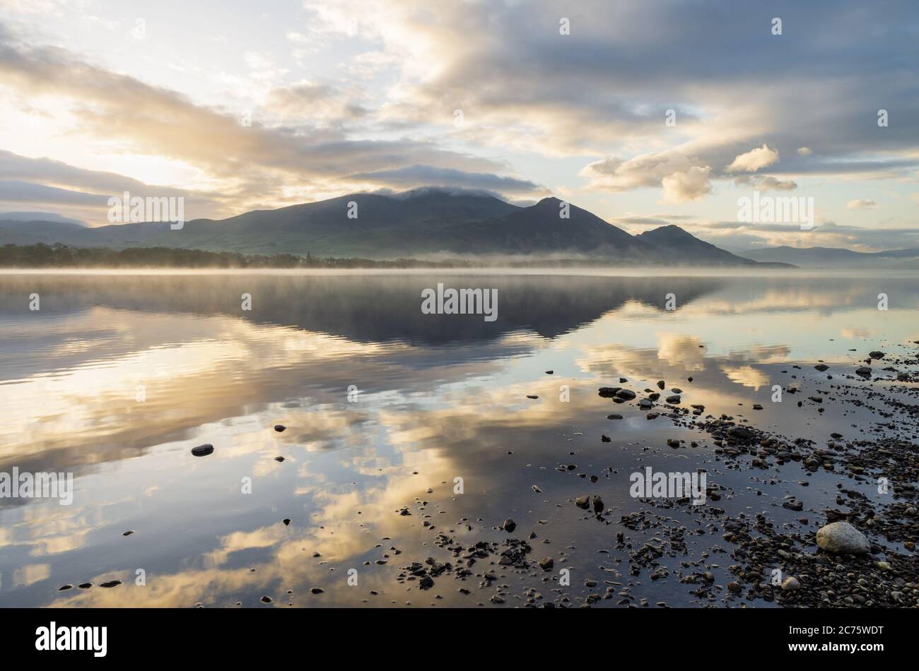 The Skiddaw Massif is bathed in early morning golden light  and reflected in the mirror calm water of Bassenthwaite Lake in the English Lake District. Stock Photo