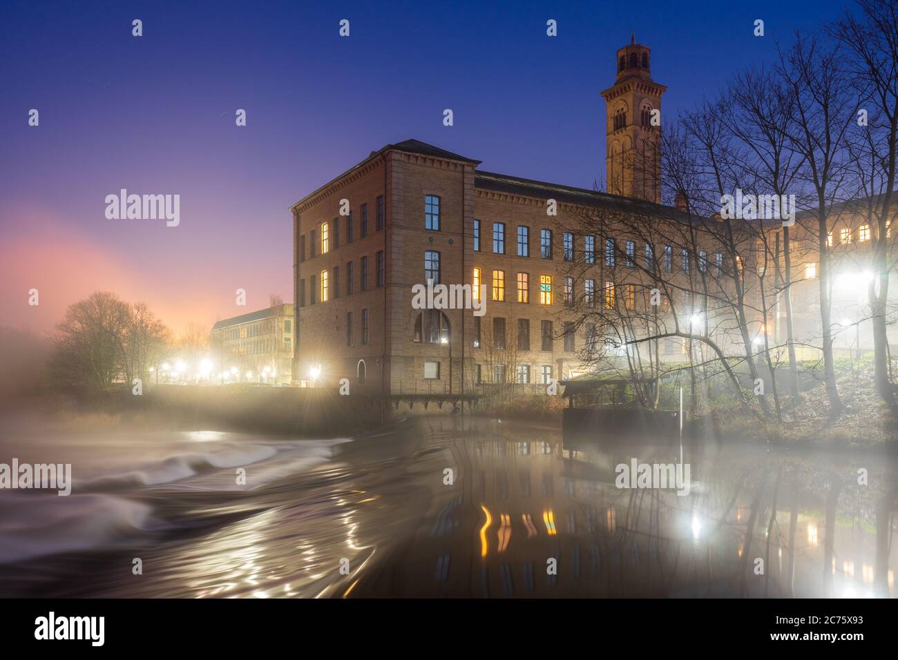 Mist forms over the River Aire on a clear winter evening and is illuminated by the lights surrounding the New Mill at the UNESCO Salts Mill site. Stock Photo