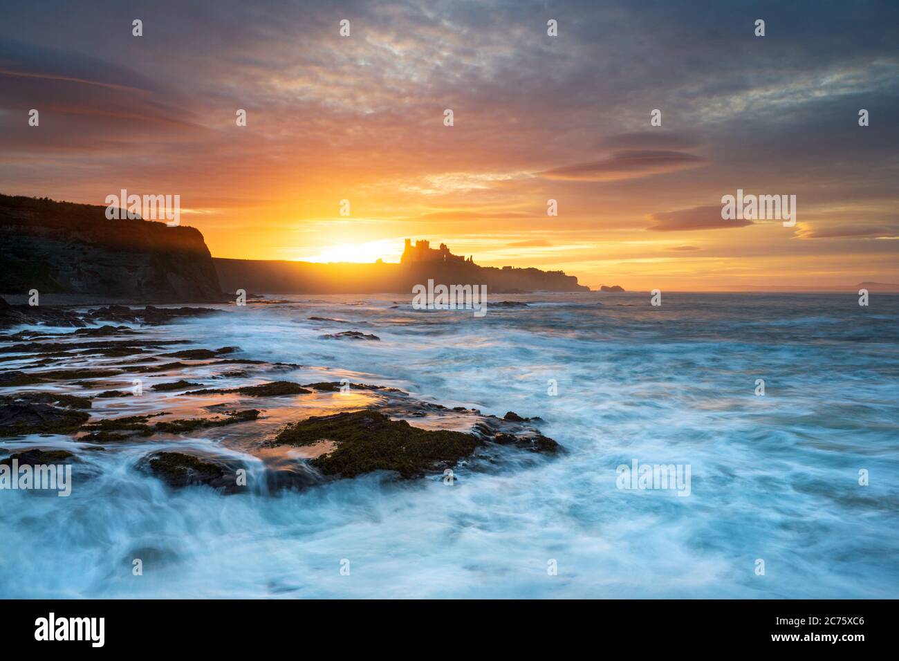 Tantallon Castle is silhouetted by the setting sun on the cliff top above Seacliff Beach, East Lothian, Scotland, as waves crash on the shore below. Stock Photo
